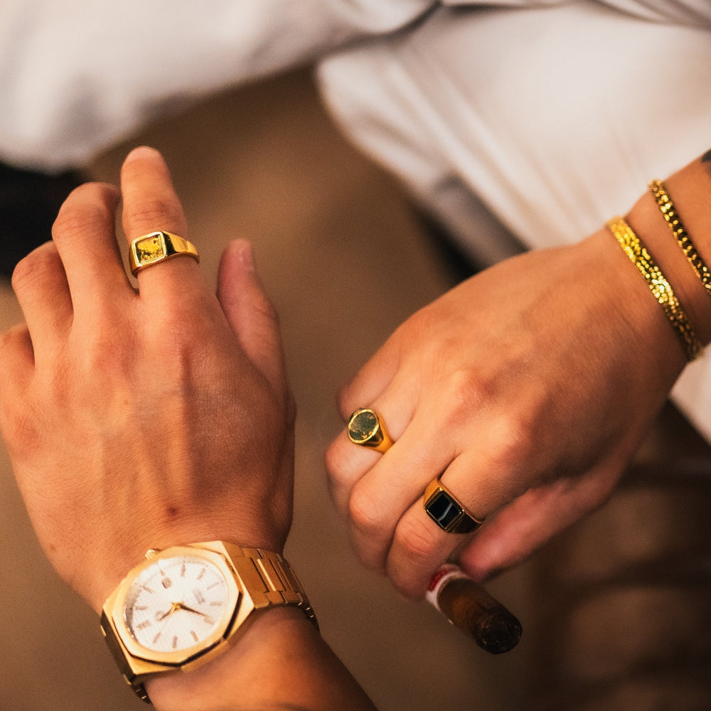 Close-up of hands wearing gold watch, rings, and bracelets on a blurred background
