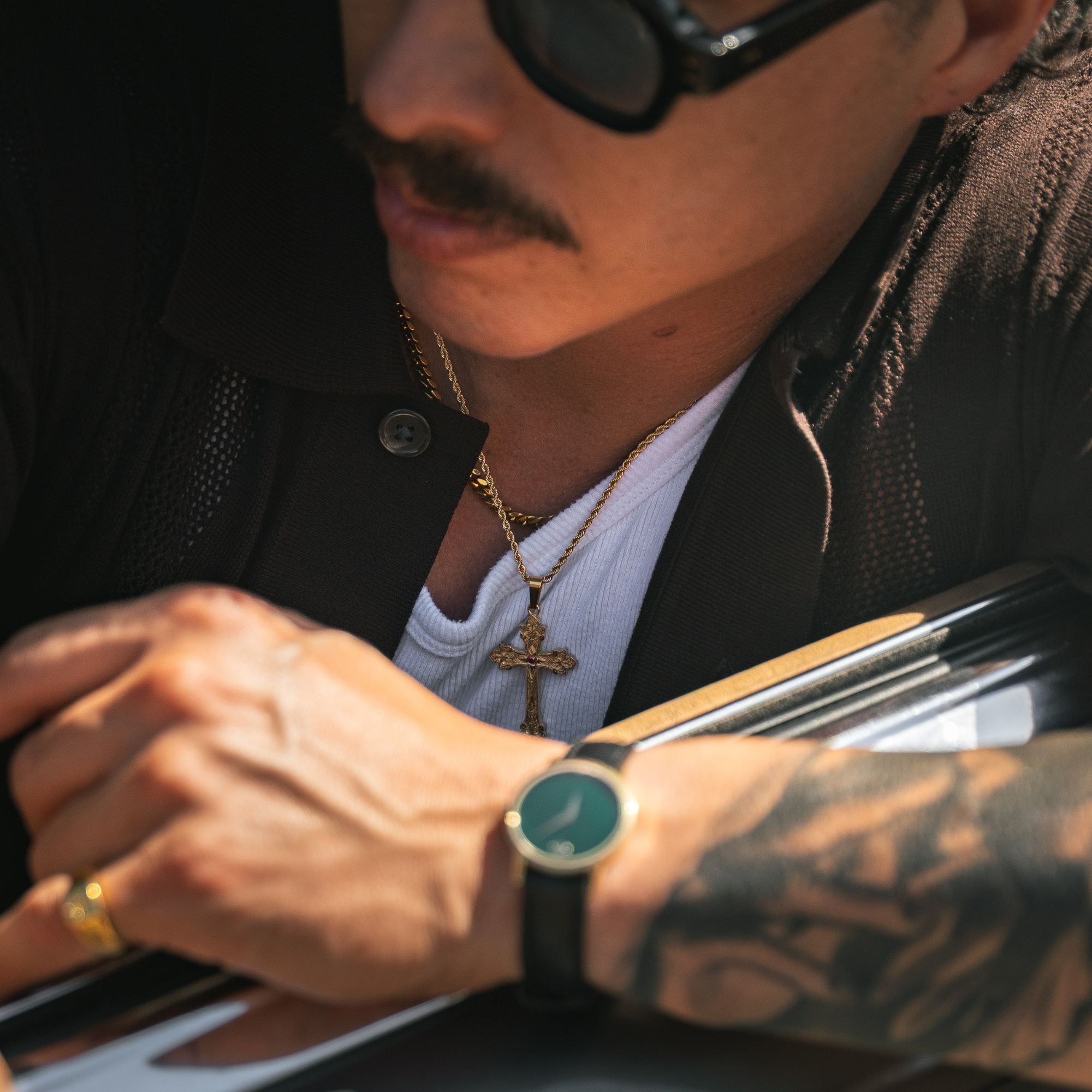 A close-up lifestyle shot of a man wearing a black leather strap watch with a round green dial and gold-tone case, styled with a white t-shirt, gold cross necklace, and sunglasses.