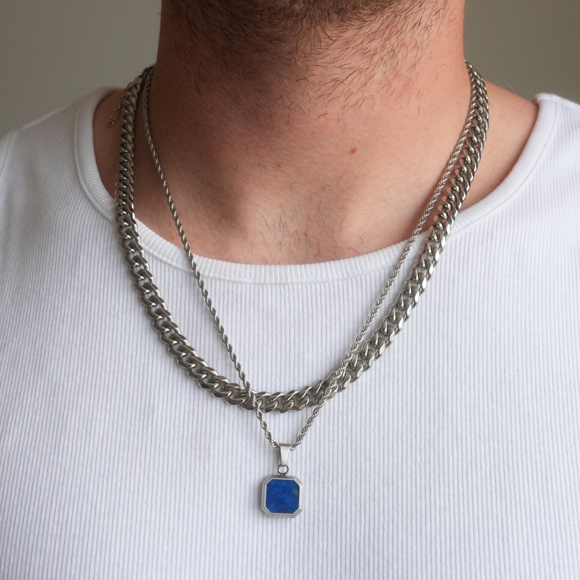 Person wearing a silver chain necklace with a blue pendant on a plain background