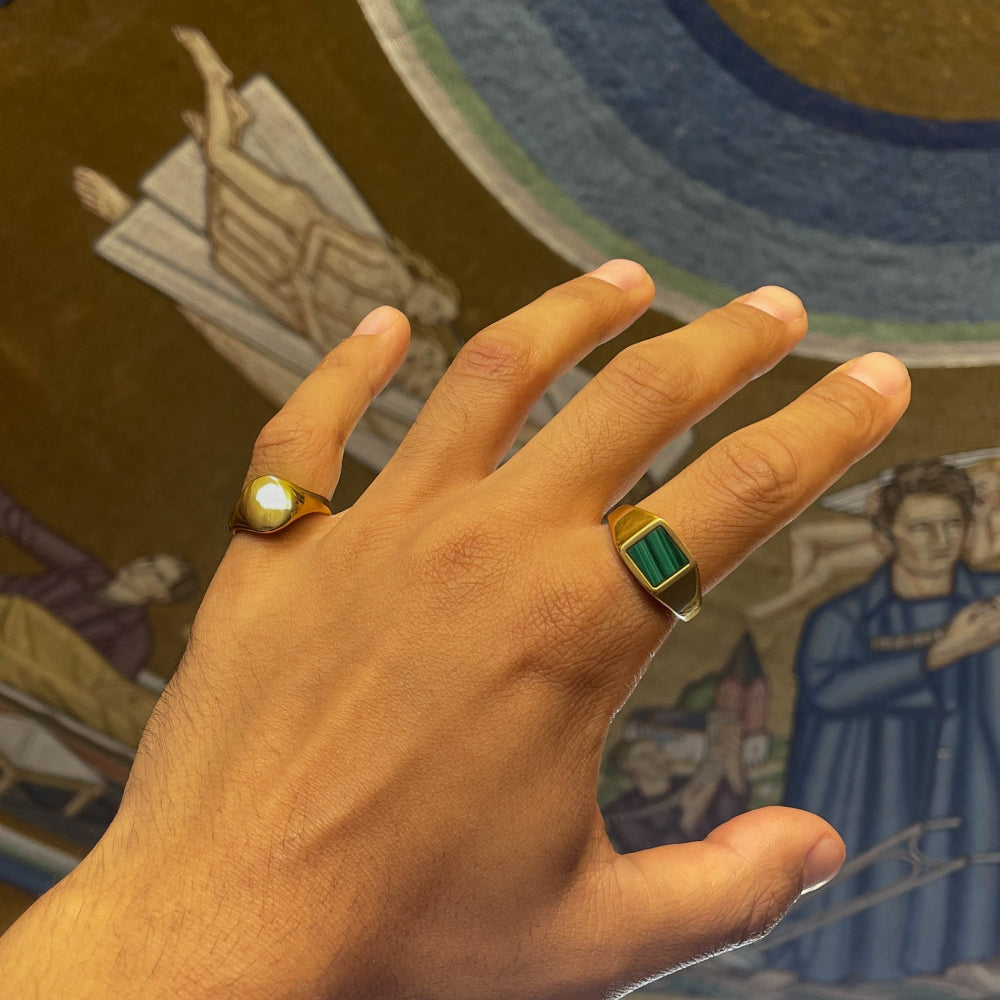 Hand wearing two gold rings with gemstones against a colorful tapestry background
