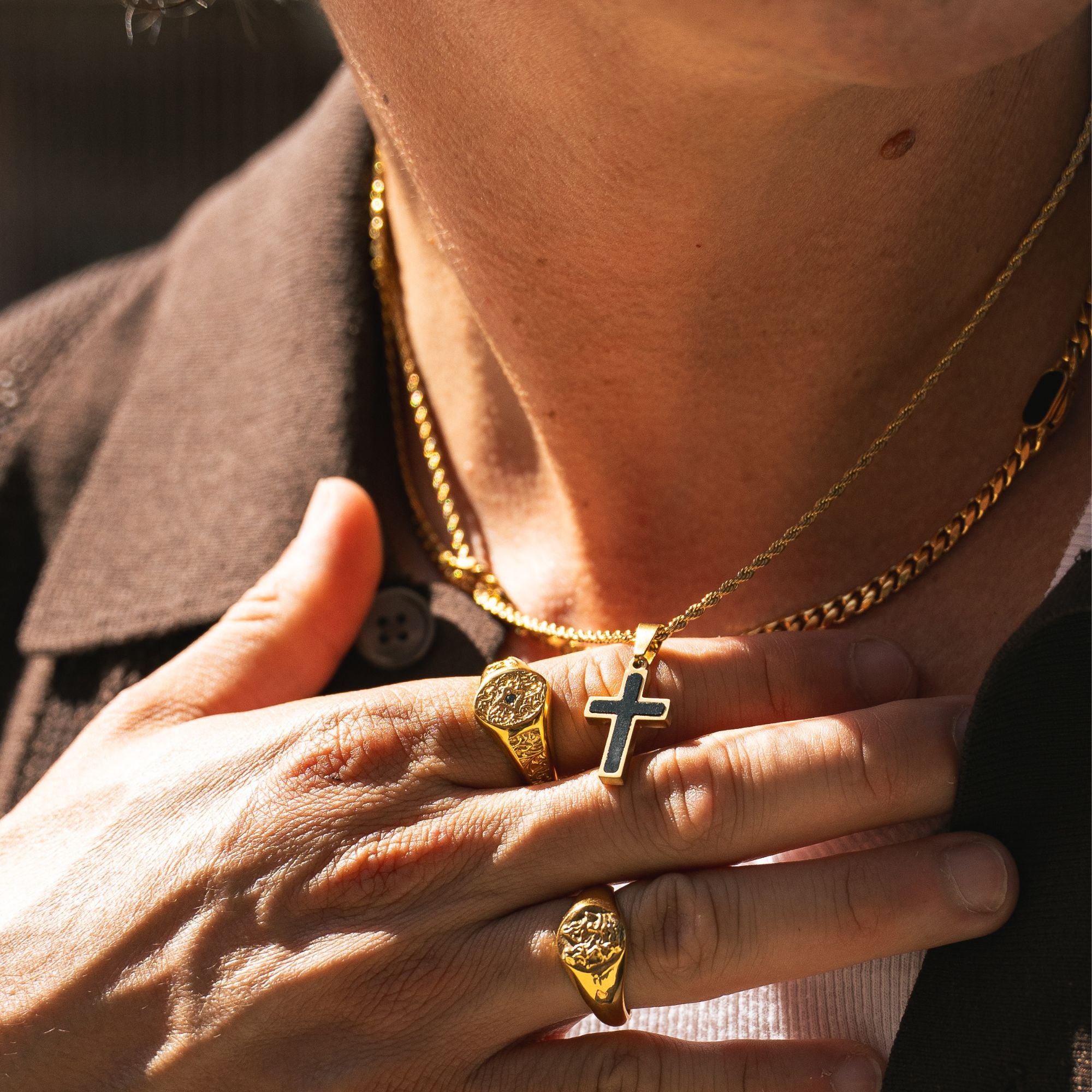 A close-up lifestyle shot of a person wearing gold jewelry, featuring a gold chain necklace with a cross pendant, gold signet rings, and a gold chain bracelet, styled with a brown collared shirt.