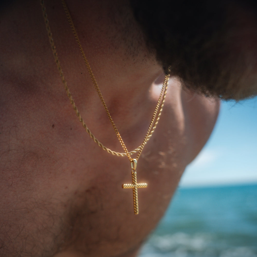 Gold necklace with a cross pendant worn by a person against a blurred beach background