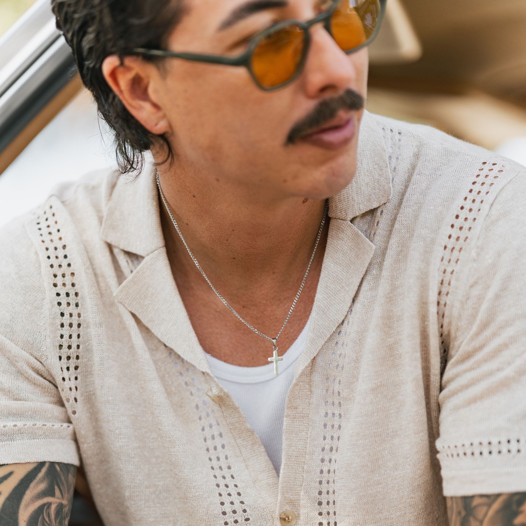 A close-up lifestyle shot of a man wearing a light beige short-sleeve linen shirt with eyelet cutouts, a white undershirt, a silver cross necklace, and orange-tinted sunglasses.