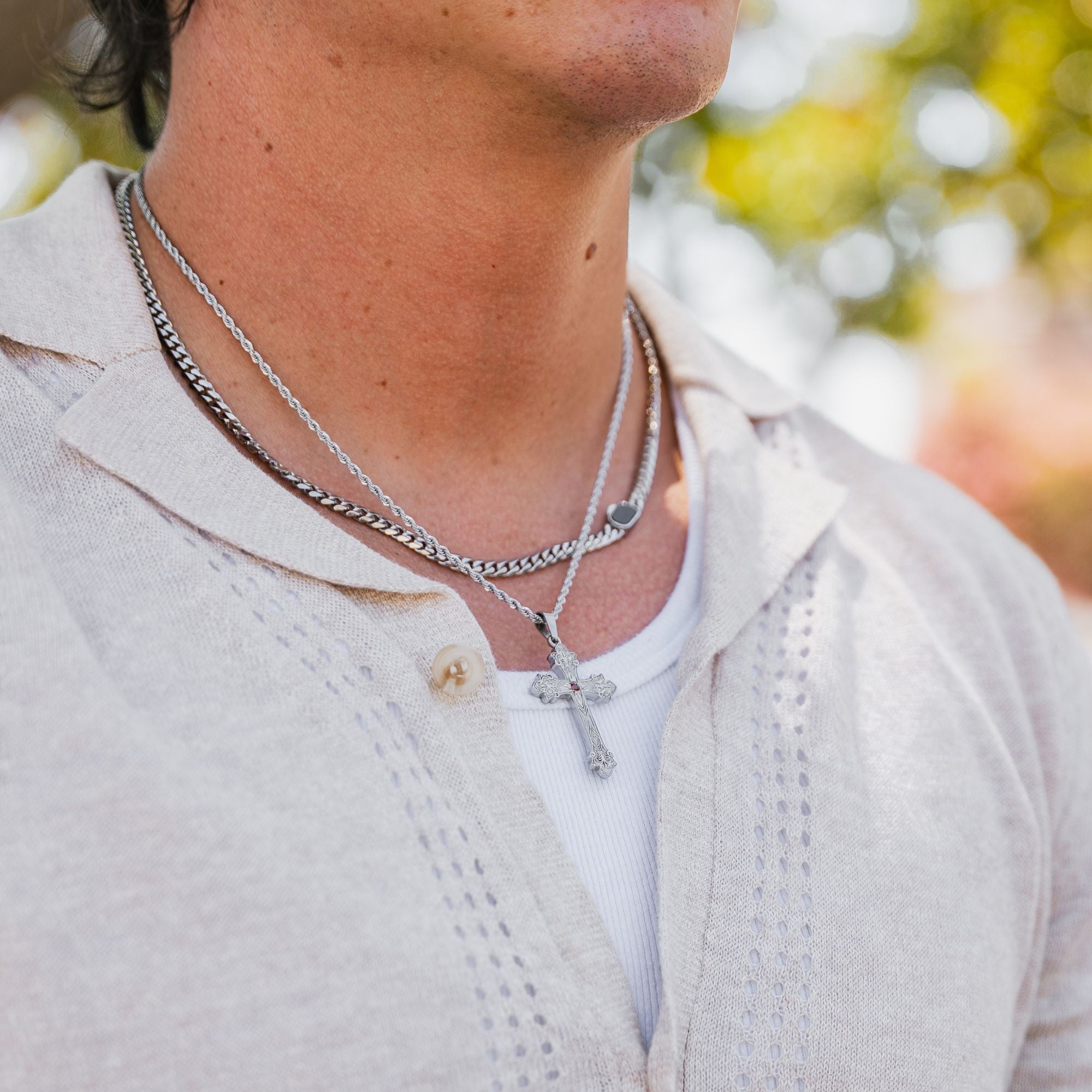 A close-up of a man's neck and chest wearing layered silver necklaces, including a chain necklace and a cross pendant, over a white t-shirt and light beige linen shirt.