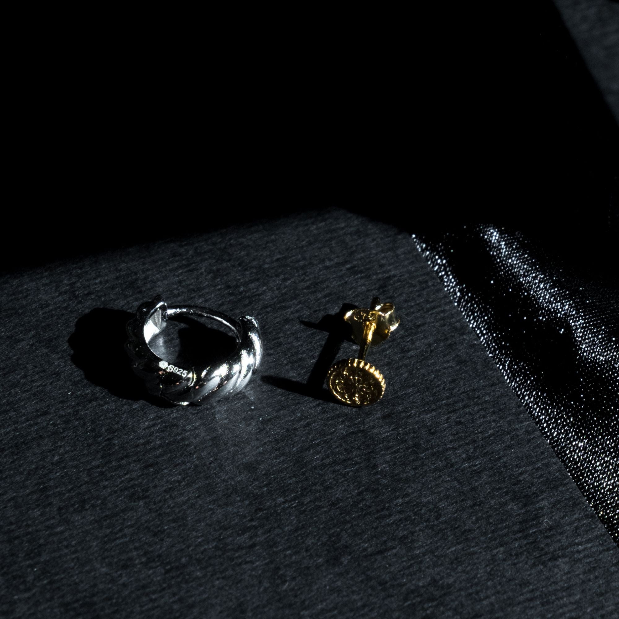 A silver twisted band ring and a gold stud earring with a round textured face, displayed on a dark textured surface with dramatic lighting.