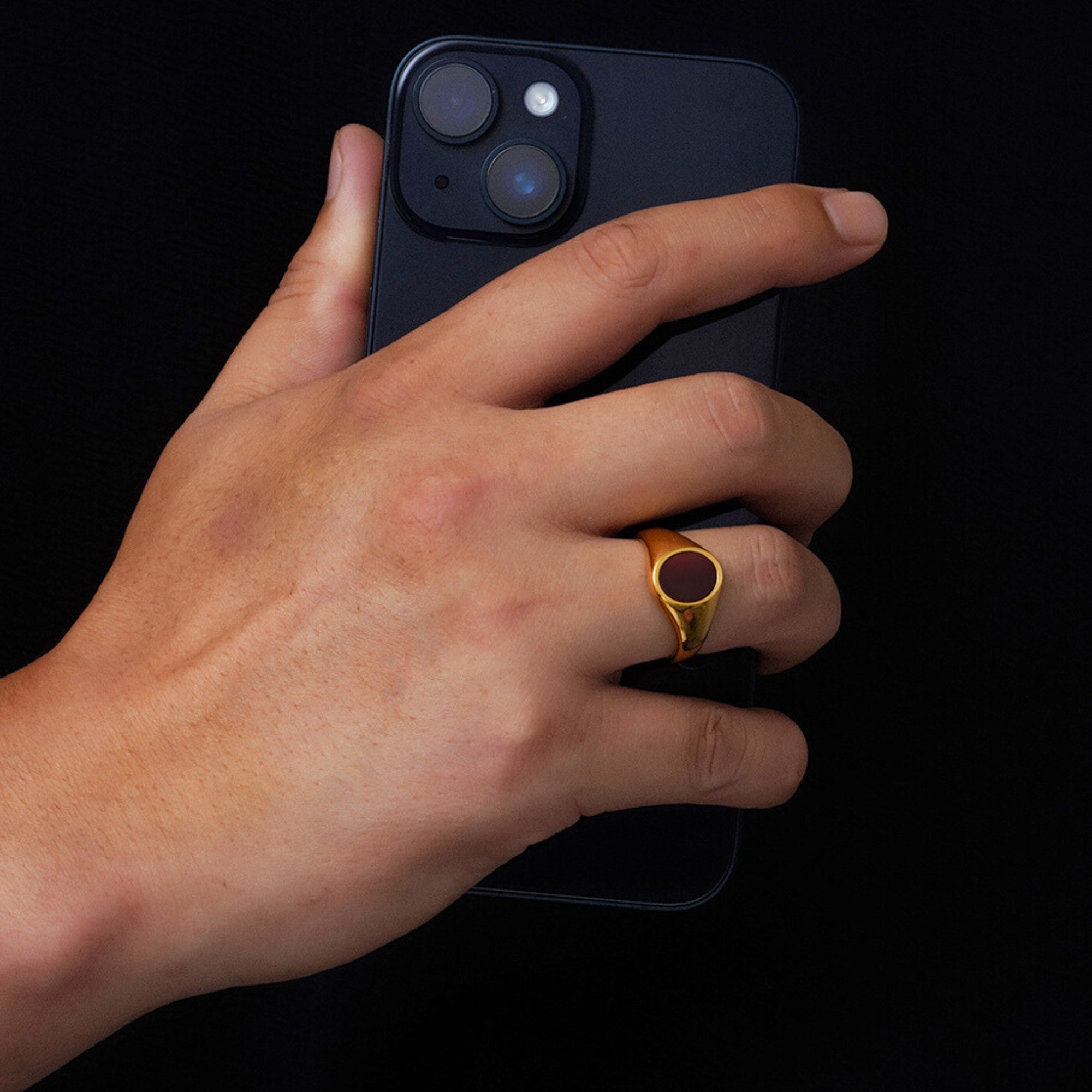 Hand holding a black smartphone with a gold ring on a black background