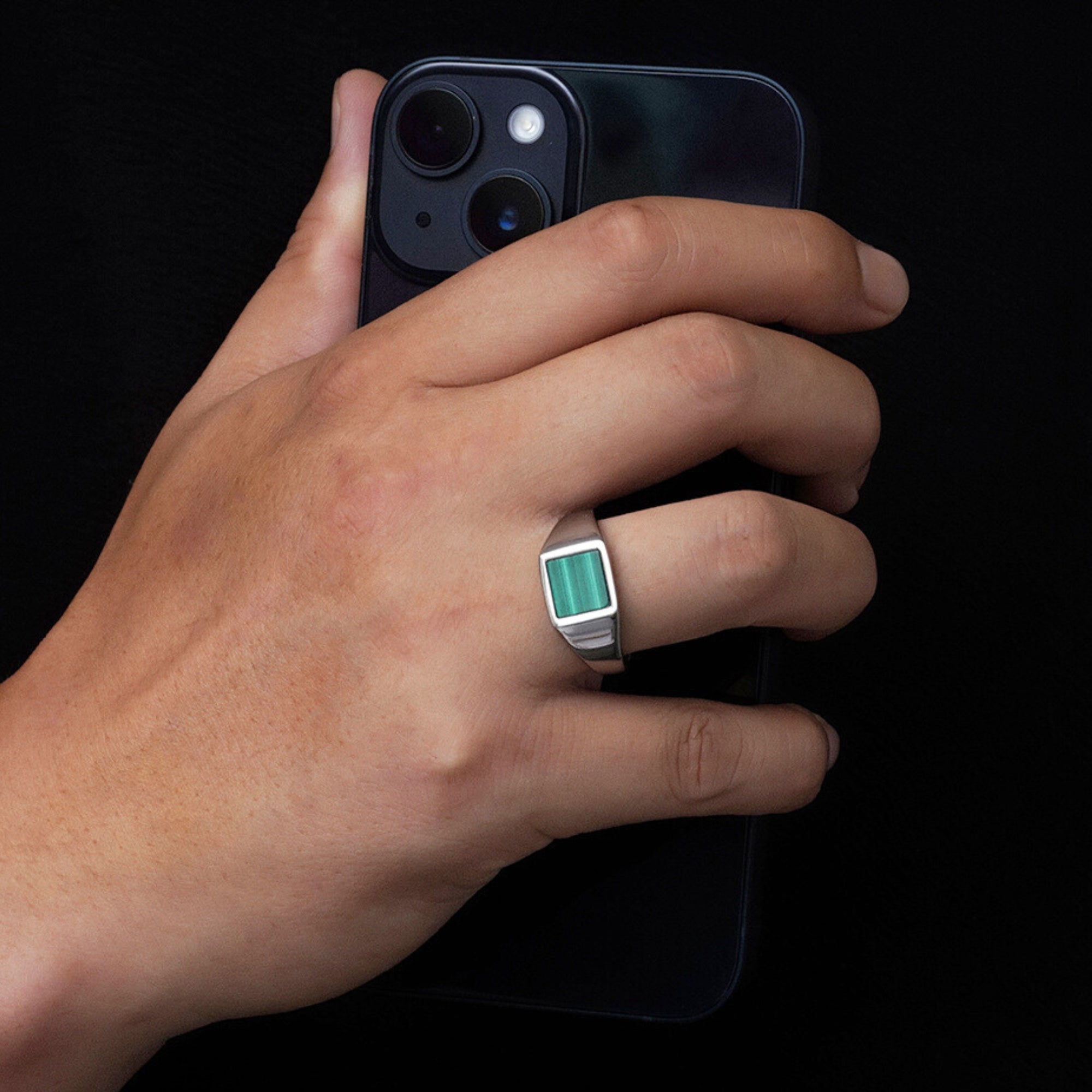 Hand holding a smartphone with a ring featuring a green gemstone on a black background