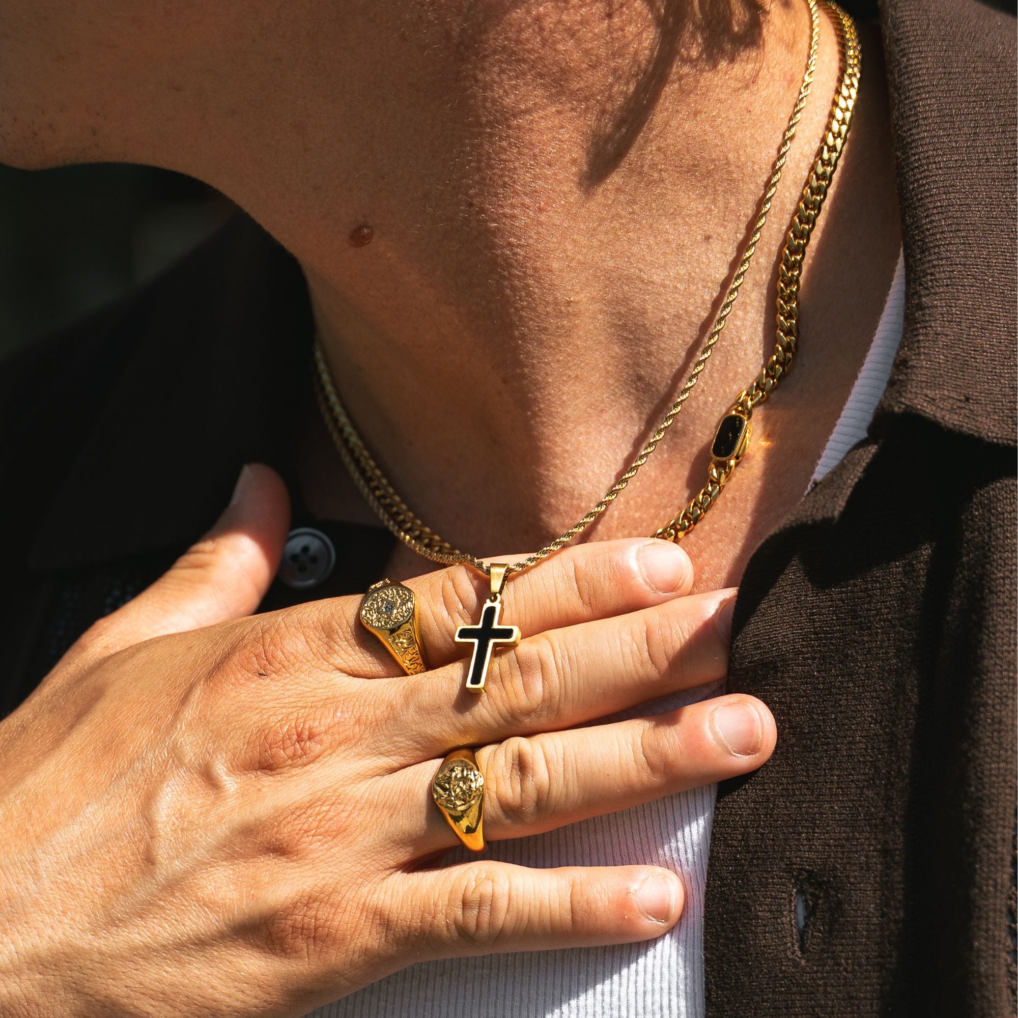 A close-up lifestyle shot of a person wearing gold jewelry, featuring a gold Cuban link chain necklace with a cross pendant, a gold rope chain necklace, and two gold signet rings on their fingers.