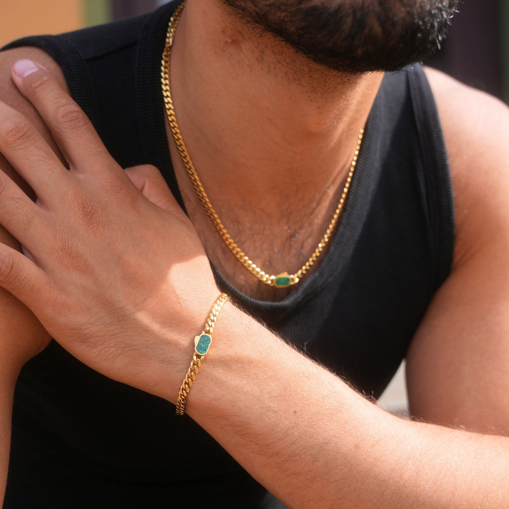 Person wearing a gold chain necklace and bracelet with green stones on a blurred background