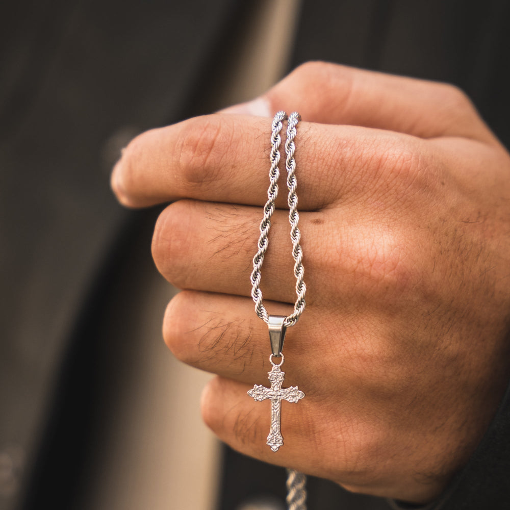 Hand holding a silver rope chain necklace with a cross pendant against a blurred background