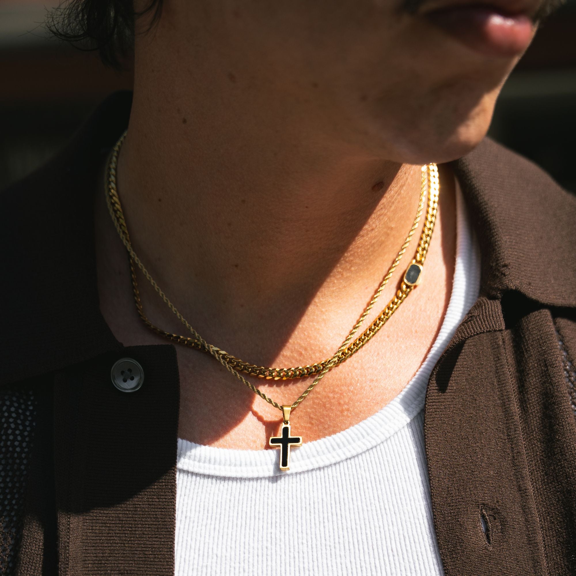 A close-up of a person's neck and lower face wearing layered gold necklaces, including a cross pendant, over a white shirt and brown jacket.