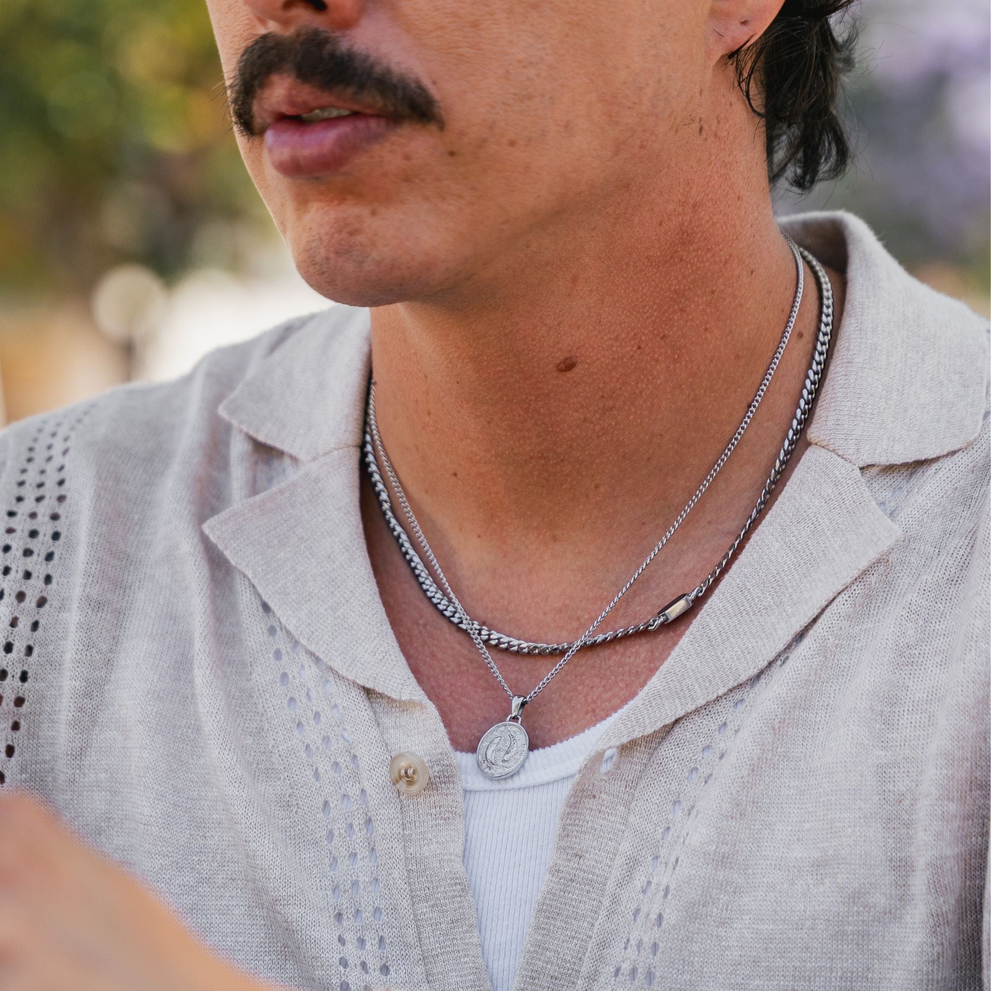 A close-up of a man wearing layered silver necklaces, including a chain necklace and a pendant necklace, over a light beige linen shirt.