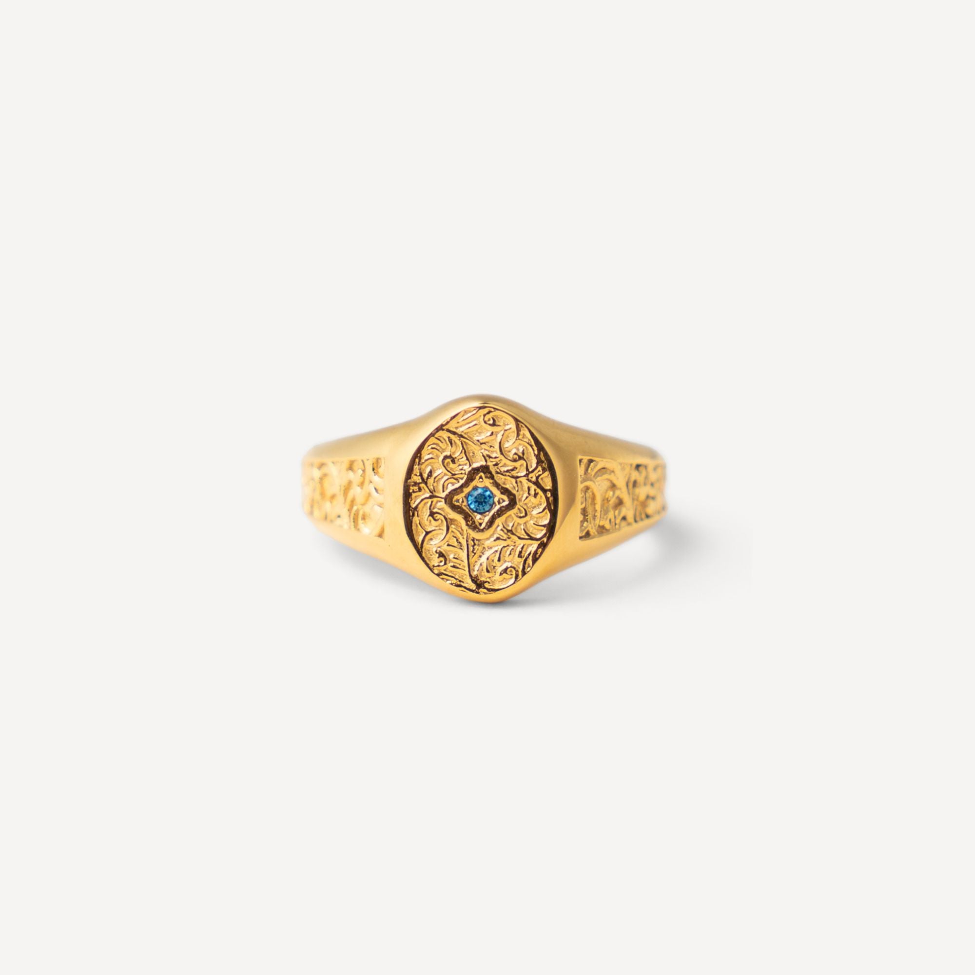 A gold signet ring with an ornate floral filigree face and a small central blue gemstone, shown on a clean off-white background.