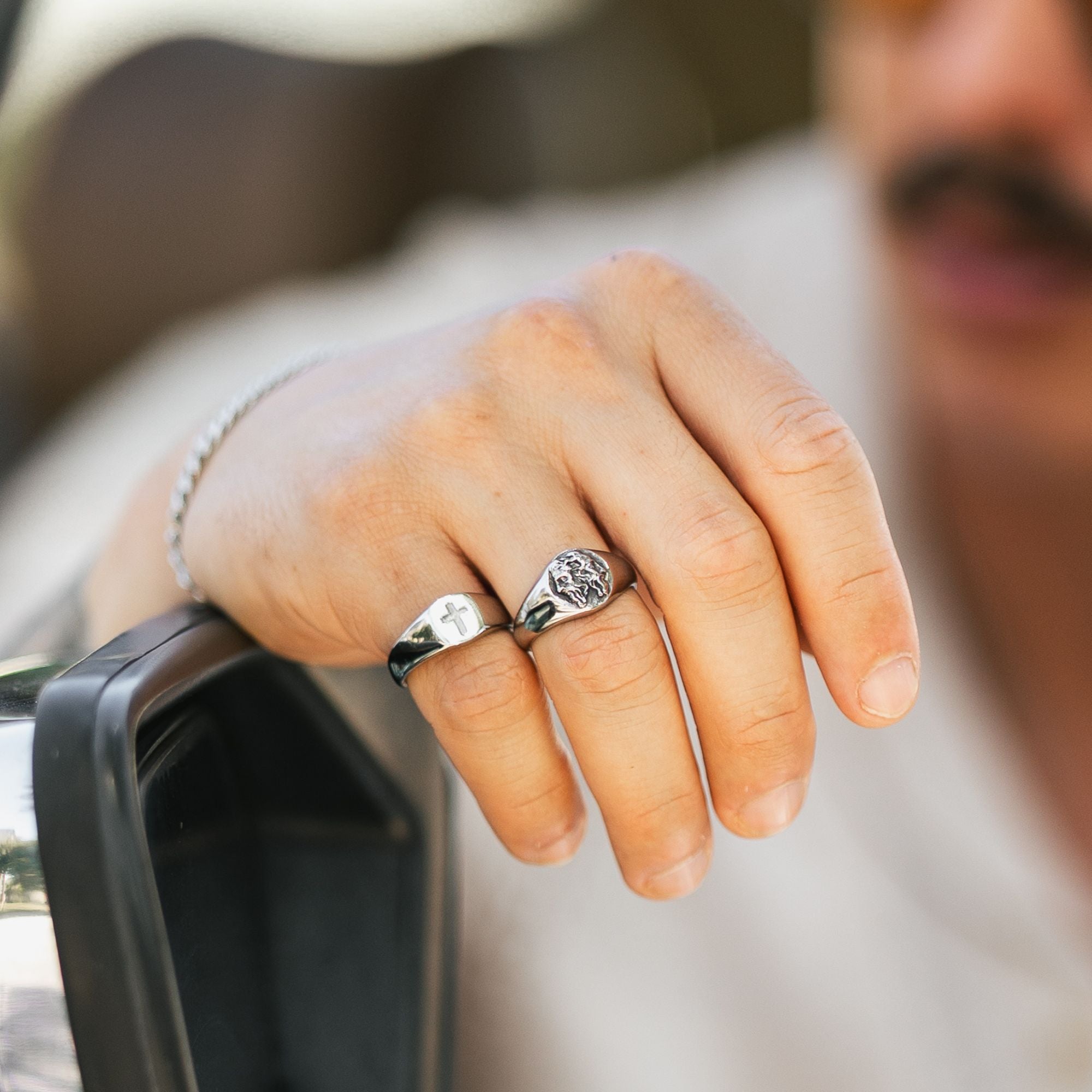 A close-up of a man's hand wearing two silver rings, one plain engraved and the other featuring a detailed emblem, with a blurred background.