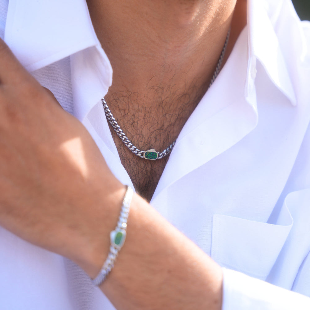 Close-up of a person wearing a silver chain necklace with a green pendant and a matching bracelet on a white shirt.