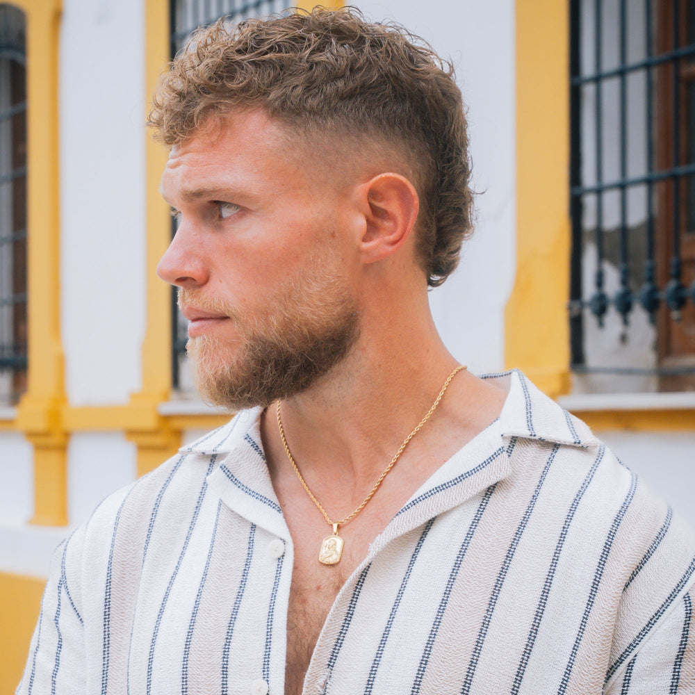Man with a beard and short haircut wearing a striped shirt and gold necklace, standing in front of a yellow building.