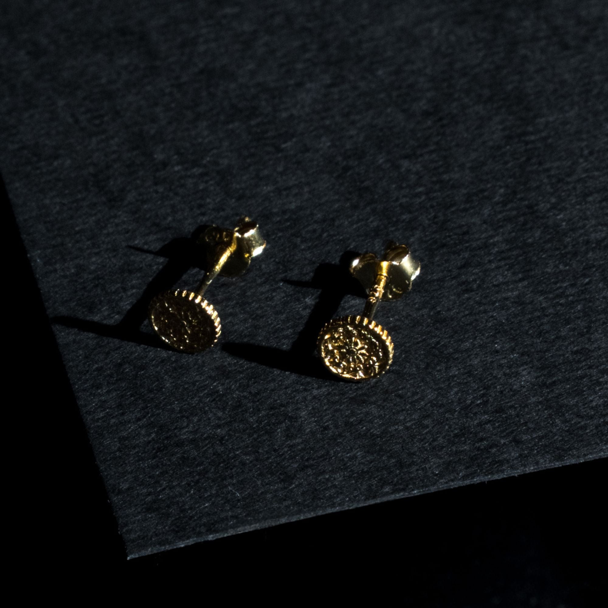 A pair of gold stud earrings with circular, textured coin-like faces, displayed on a dark textured surface with dramatic shadows.