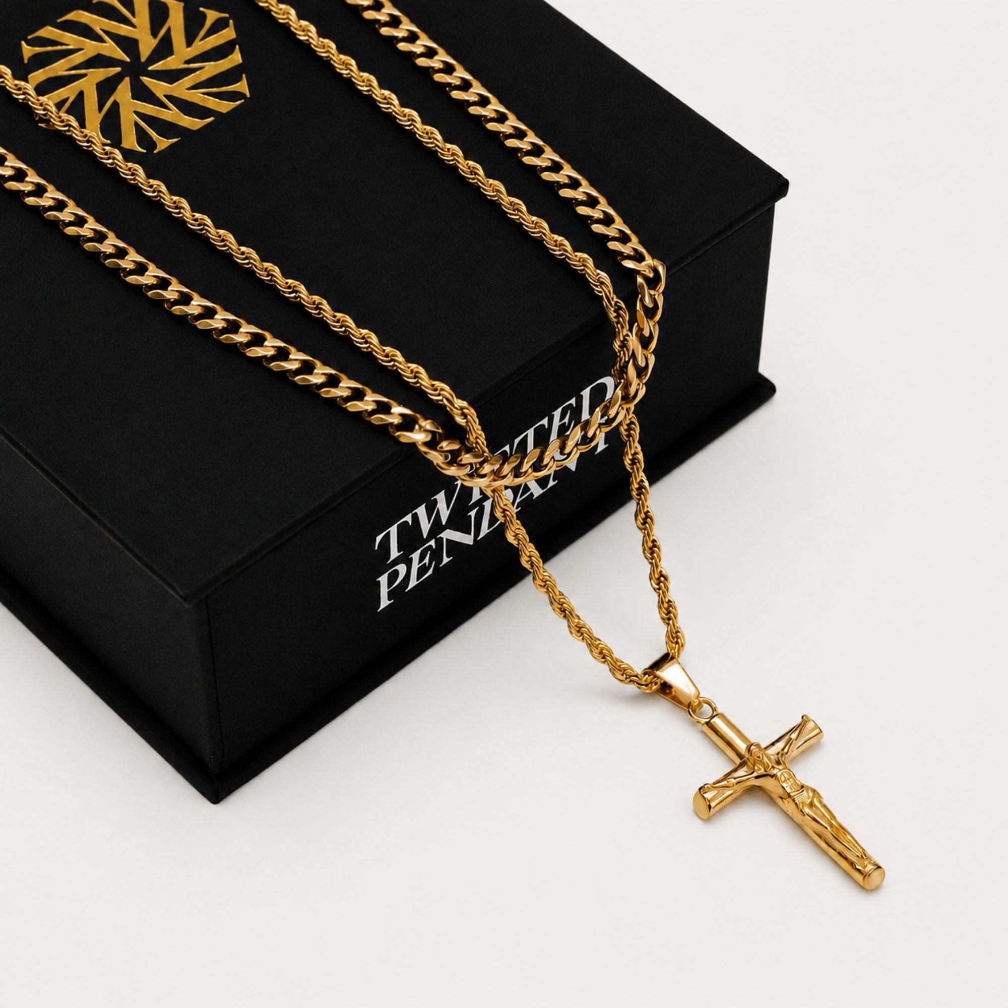 A gold-tone crucifix pendant necklace with a thick curb chain draped over a black presentation box. The box has white text reading 'TWISTED PENIS' and a gold geometric logo on the lid.