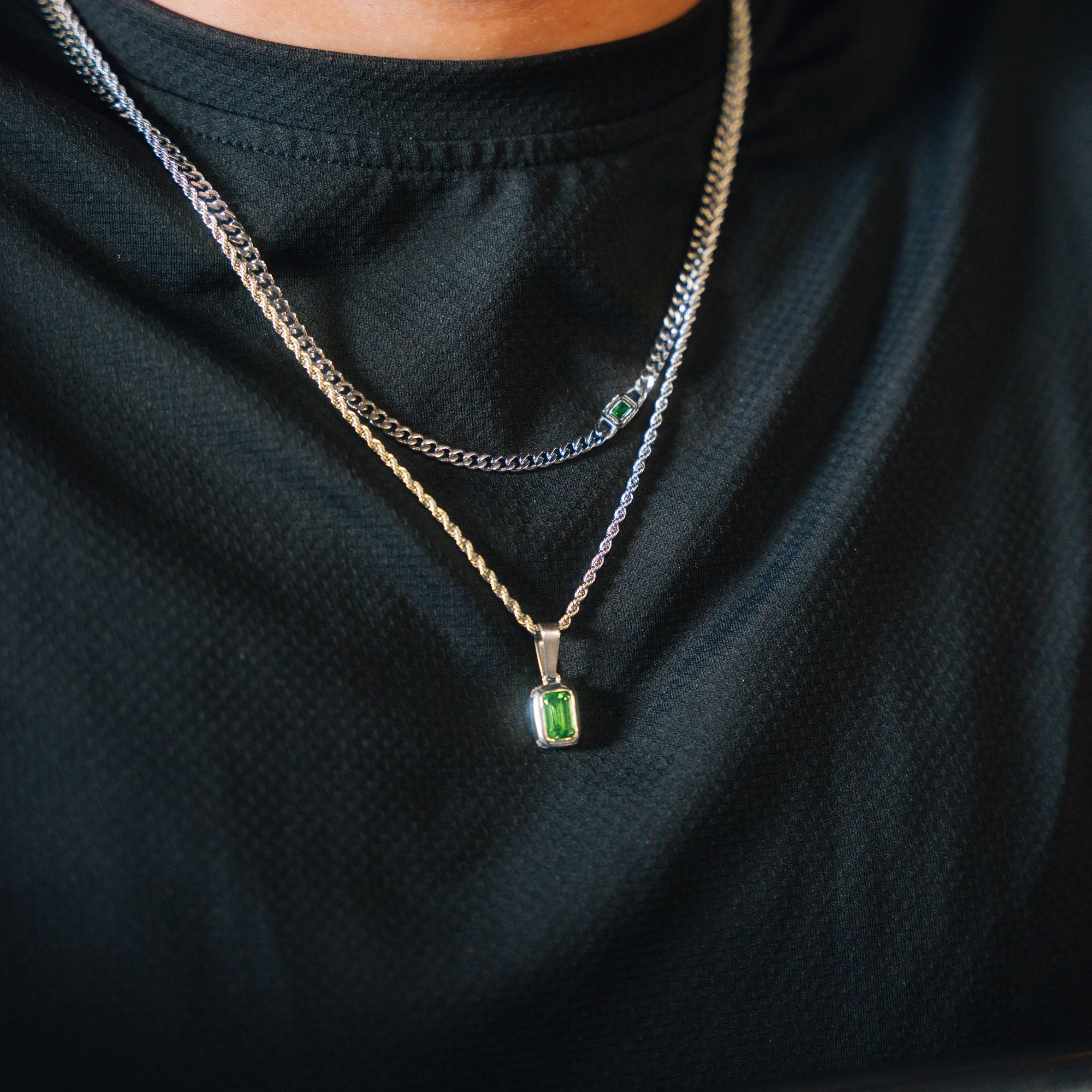 A close-up of a person wearing a layered necklace set, featuring a silver chain with a rectangular green gemstone pendant and a silver chain with a small green stone accent, over a black textured shirt.
