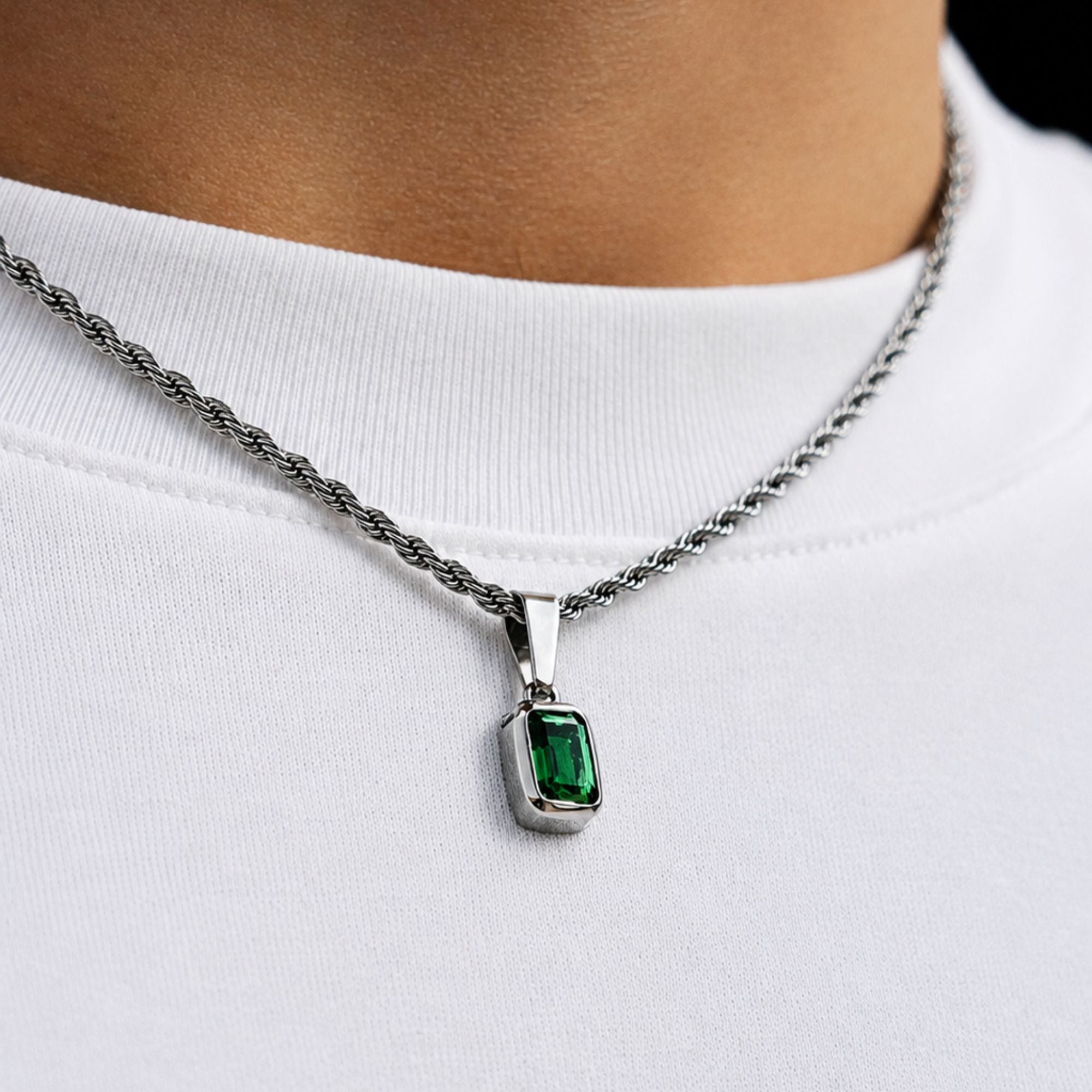 A silver-toned rope chain necklace with a rectangular green gemstone pendant, worn on a person's neck over a white t-shirt.