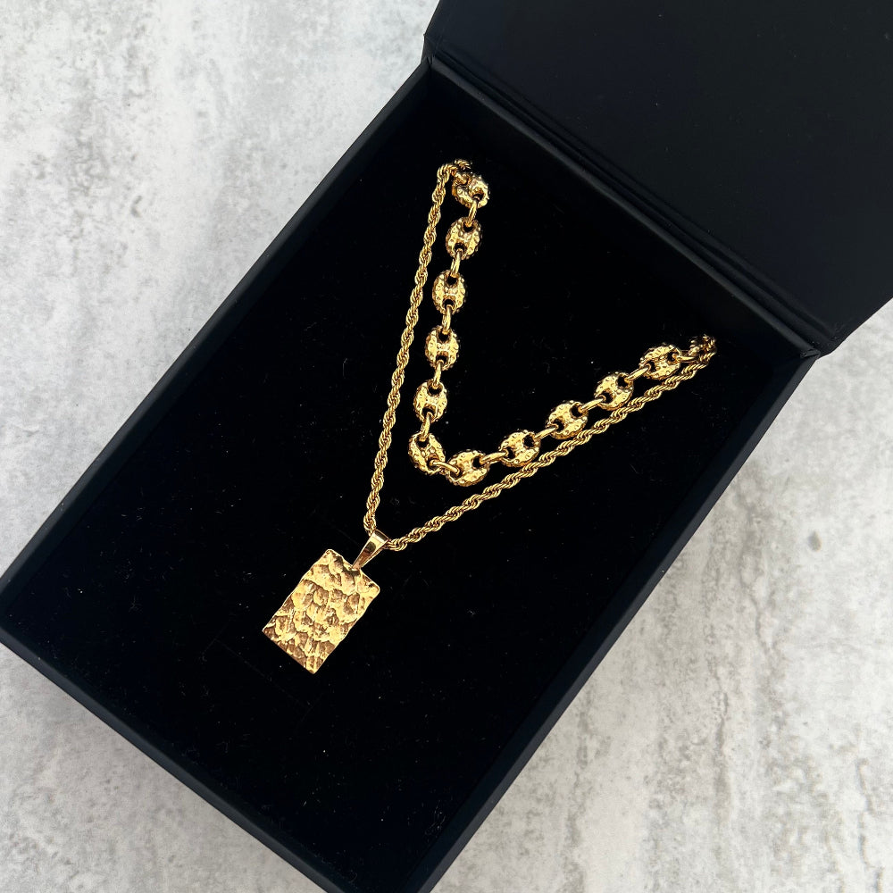 Gold necklace with a rectangular pendant on a black box against a light gray background