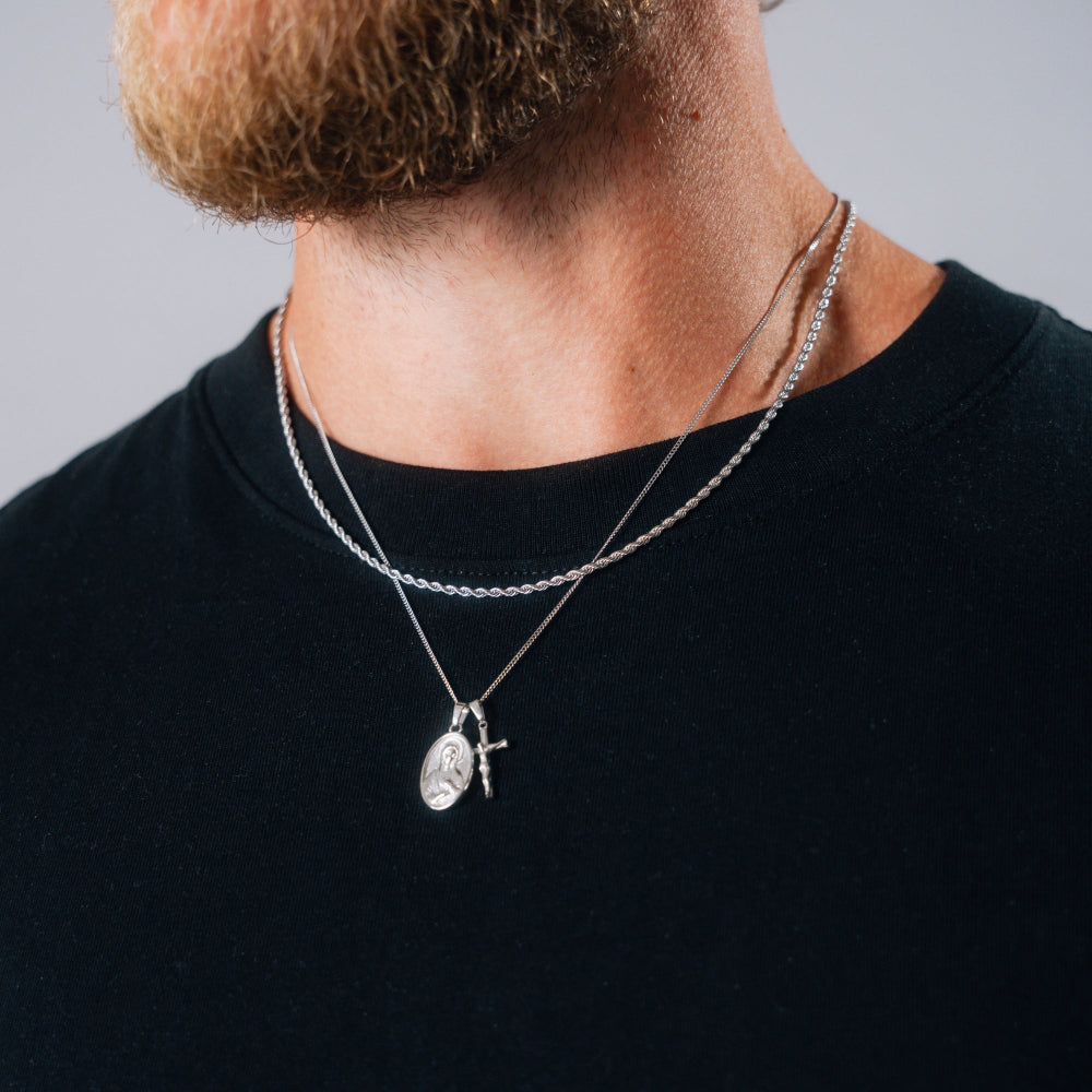 Man wearing a black shirt with two silver necklaces featuring pendants against a gray background