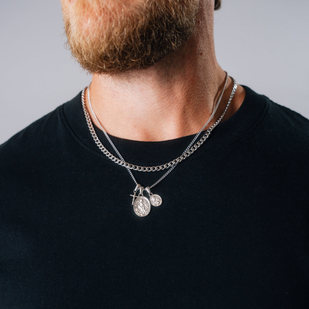 Man wearing a black shirt with a silver necklace featuring two pendants against a neutral background