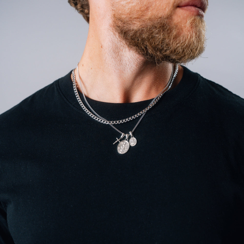 Man wearing a black shirt with a silver chain necklace featuring two round pendants on a gray background