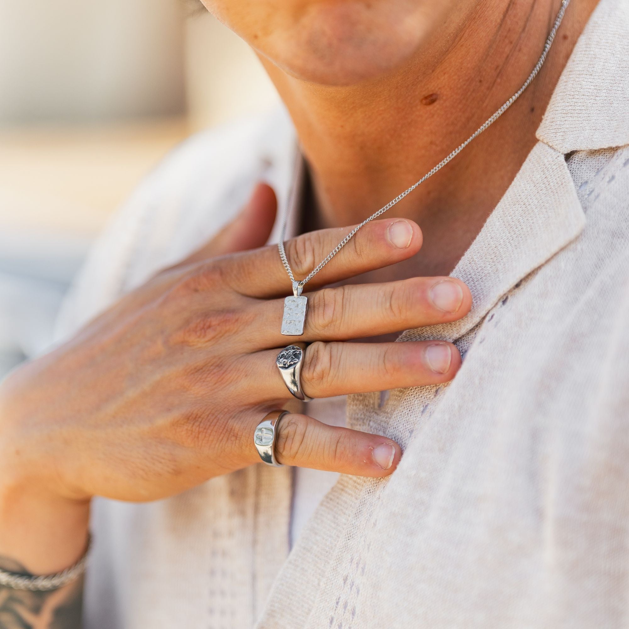 A close-up lifestyle shot of a person wearing silver rings and a silver pendant necklace, wearing a light beige linen shirt.