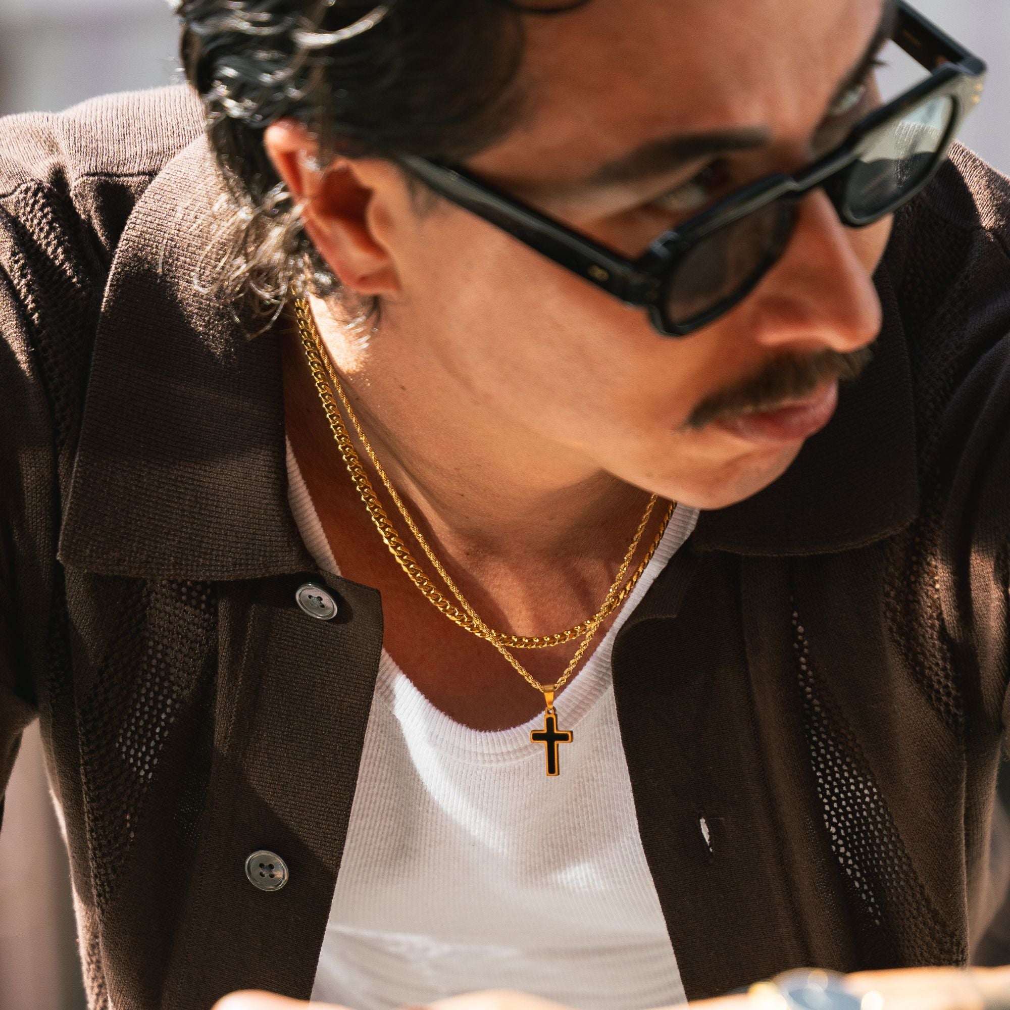 A close-up lifestyle shot of a man wearing a gold chain necklace with a cross pendant, styled with black sunglasses, a white t-shirt, and a dark brown shirt.