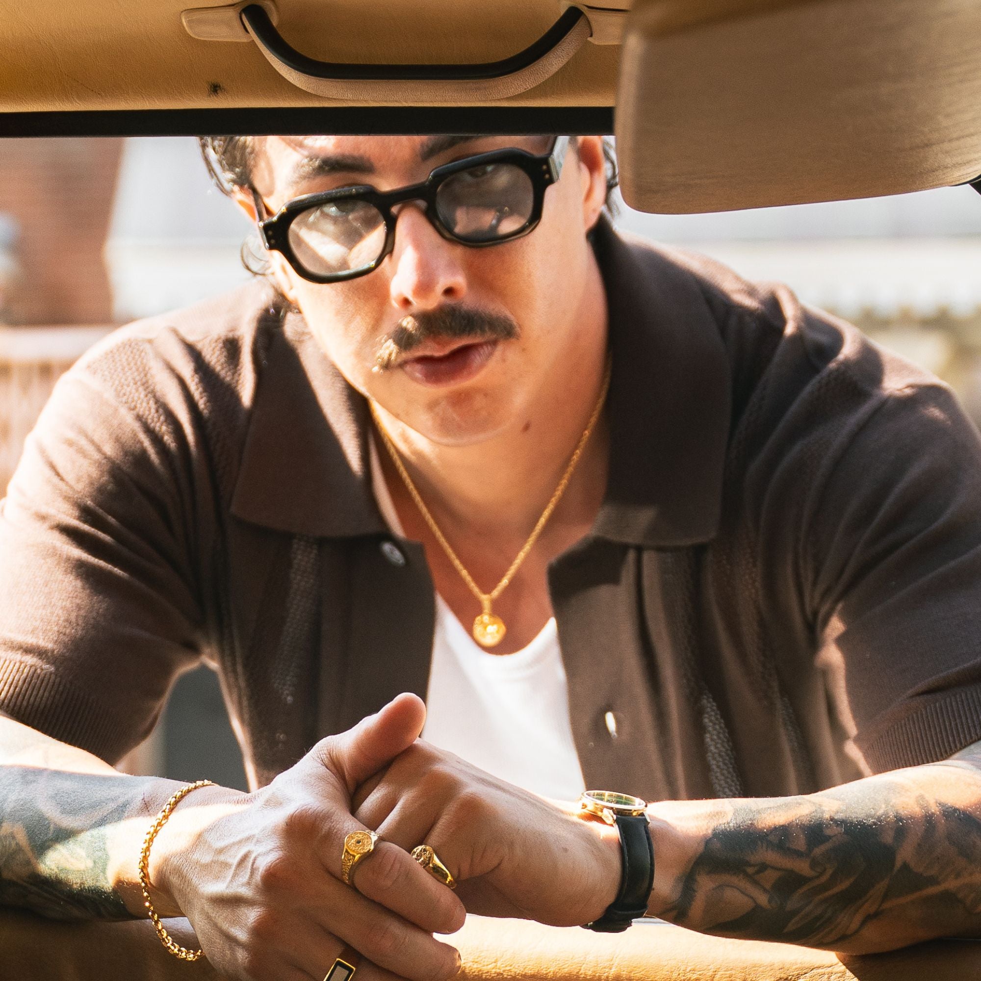 A man with a mustache and black sunglasses leans over a car windshield, wearing a dark brown knit polo shirt over a white undershirt, along with gold jewelry including a necklace, rings, and a watch.