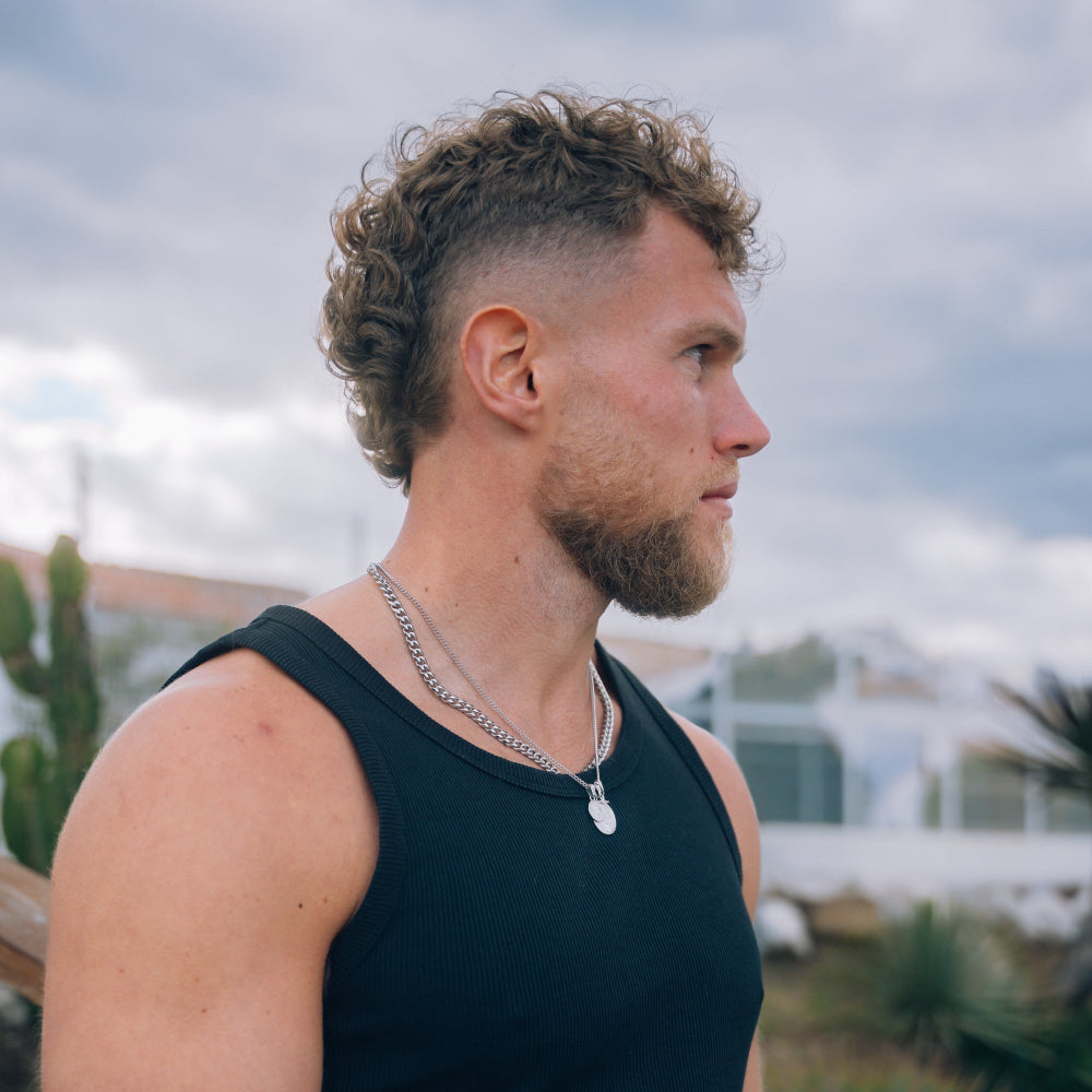 Man with a beard and curly hair wearing a black tank top and silver necklace, standing outdoors.