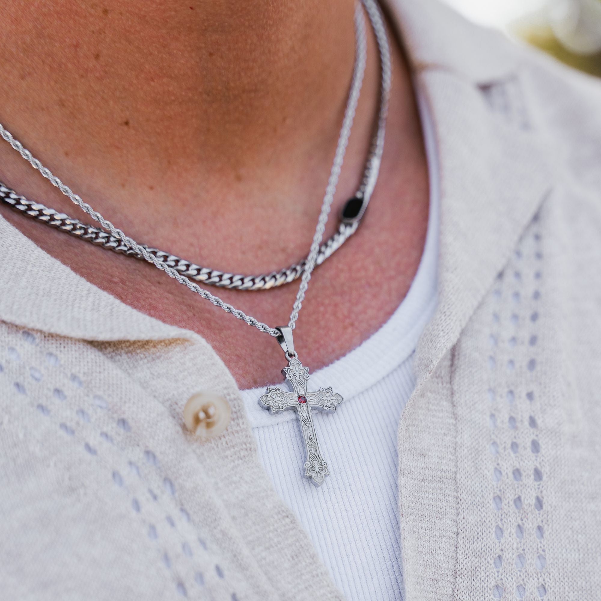 A close-up of a person wearing layered necklaces, including a silver cross pendant with a small red stone over a white shirt and a light beige knit cardigan.