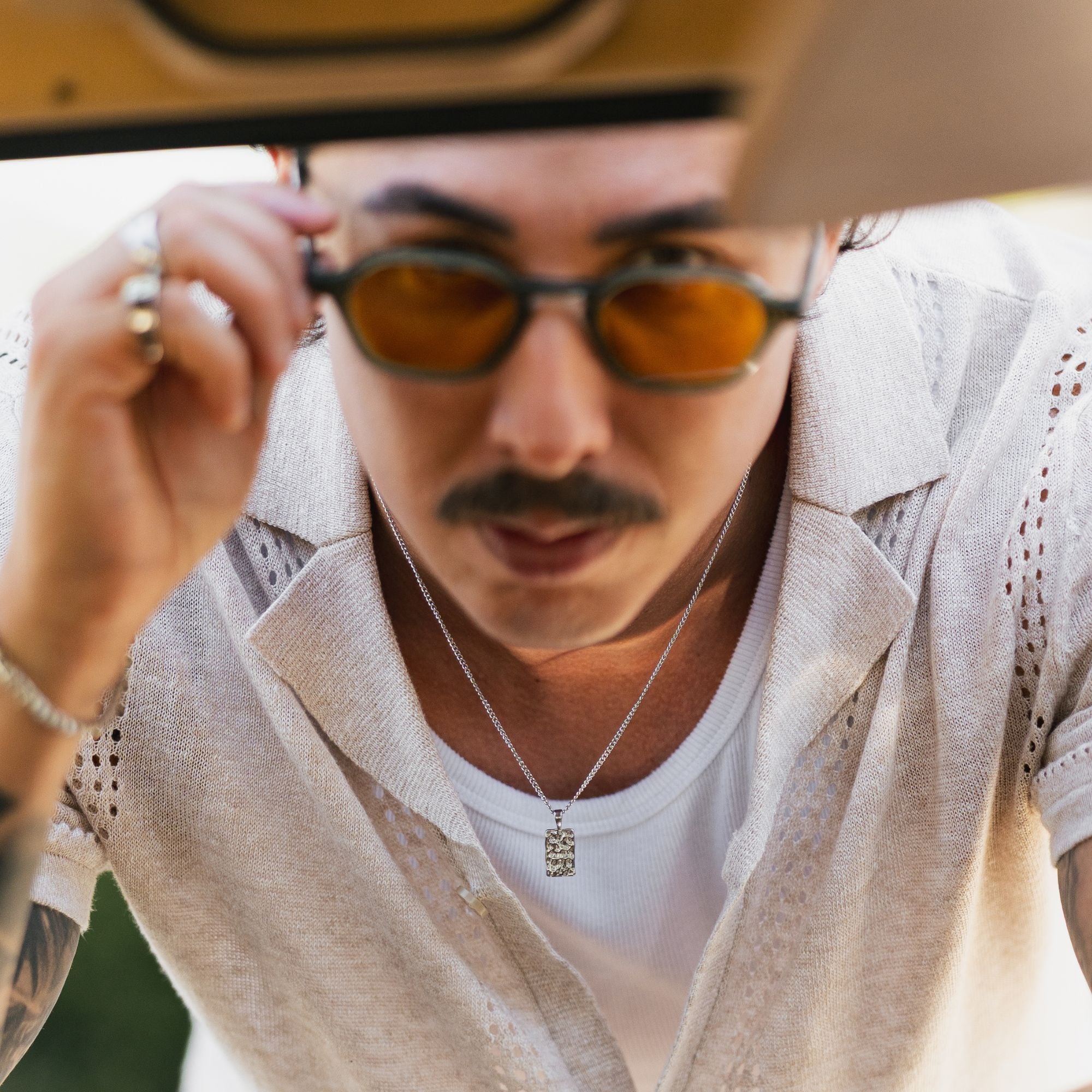 A close-up lifestyle shot of a man wearing round dark sunglasses with orange-tinted lenses, a white t-shirt, a beige textured linen shirt, and a silver pendant necklace.