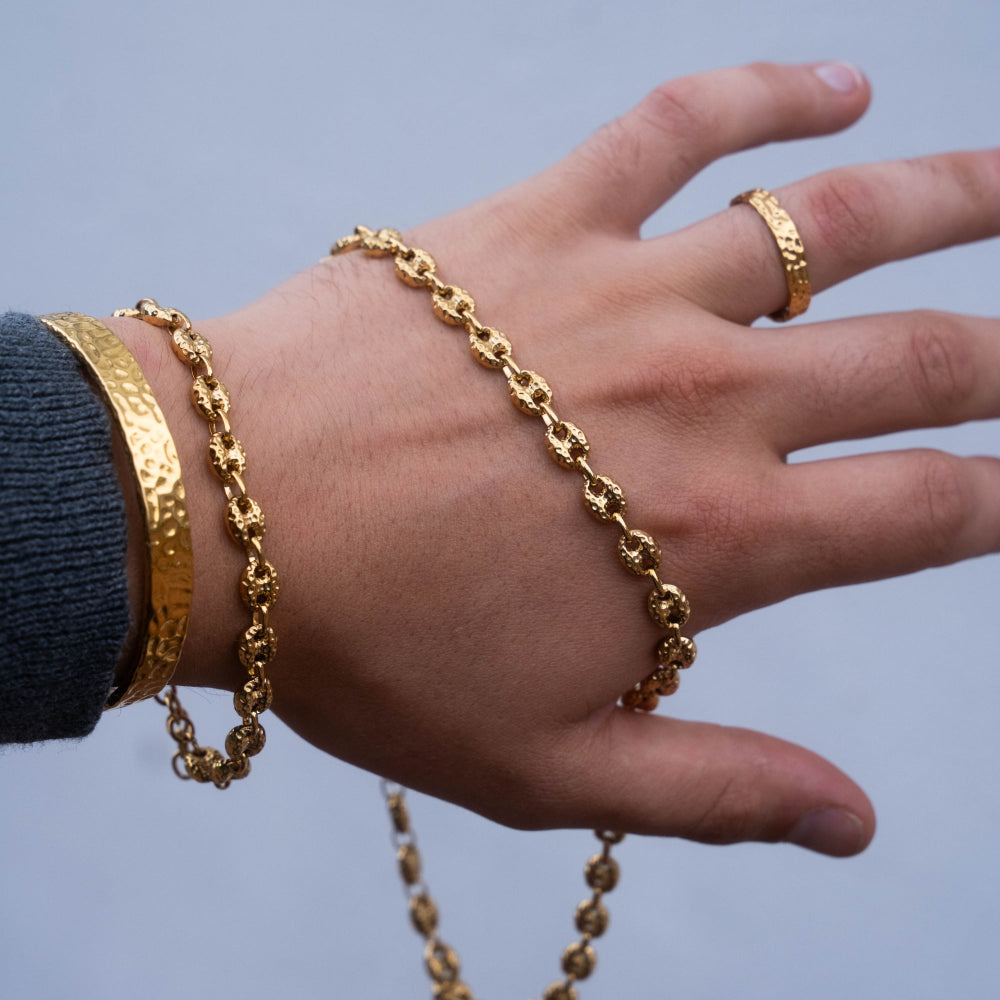 Hand wearing gold jewelry including a bracelet, ring, and bangle against a neutral background
