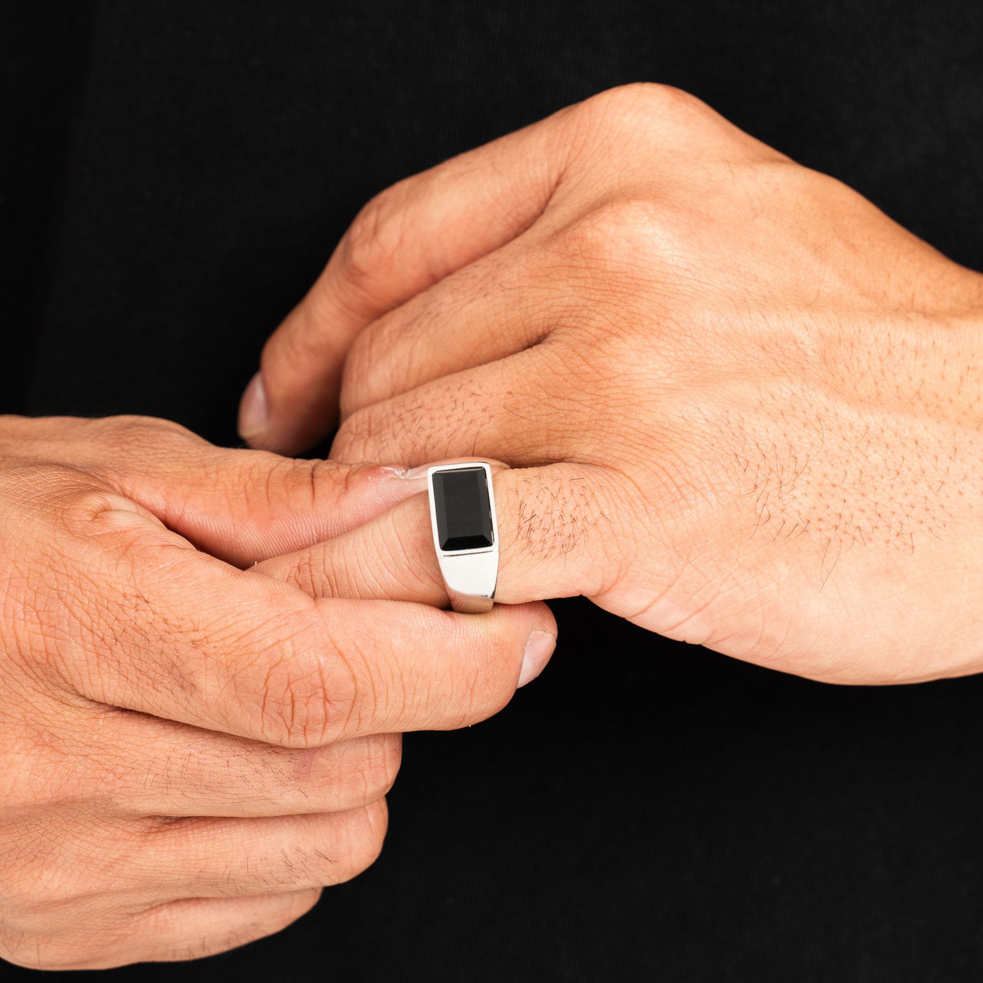 A silver-toned signet ring with a rectangular black inlay, shown being worn on a person's finger against a solid black background.