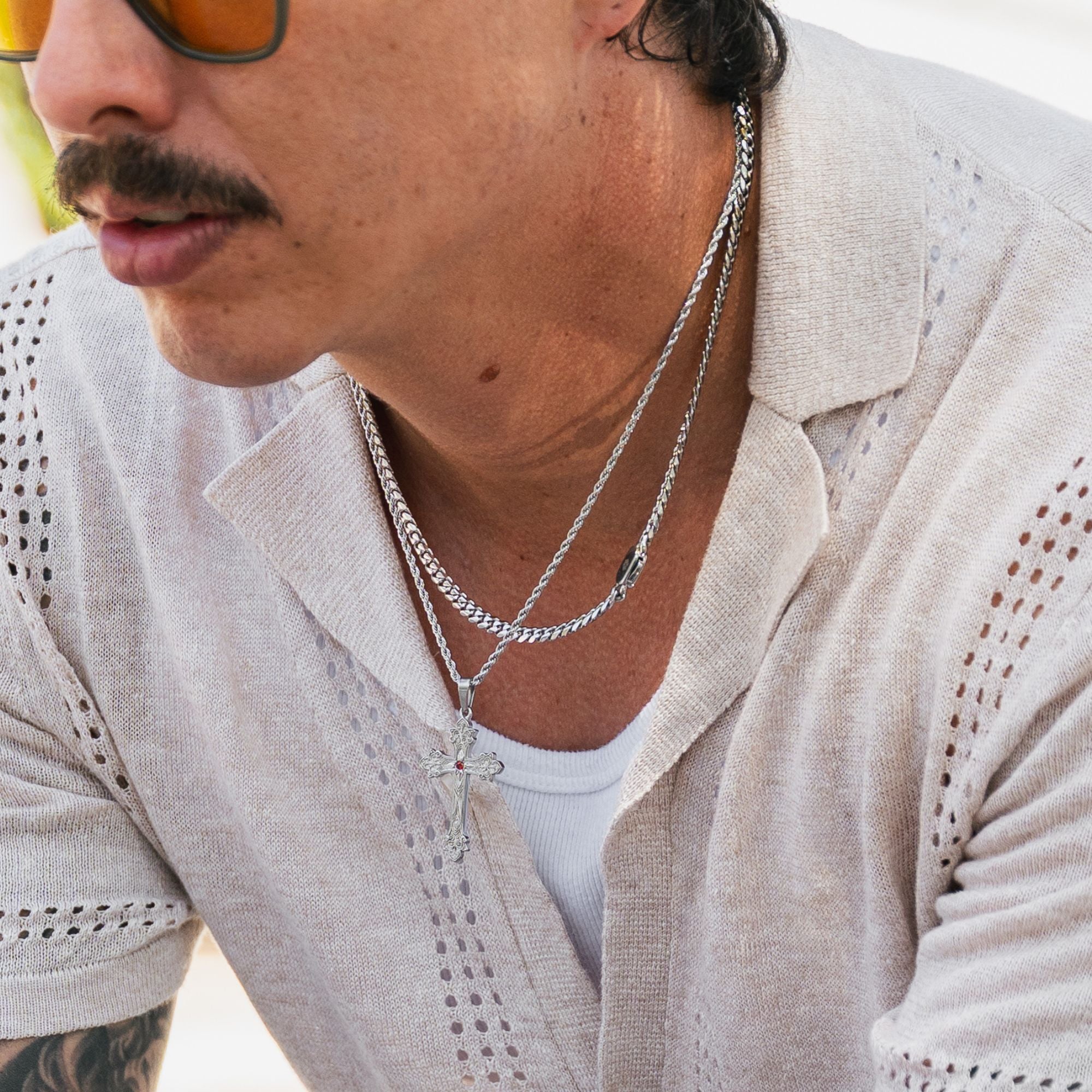 A close-up lifestyle shot of a man wearing a silver chain necklace with a cross pendant over a white ribbed undershirt and a light beige knit shirt.