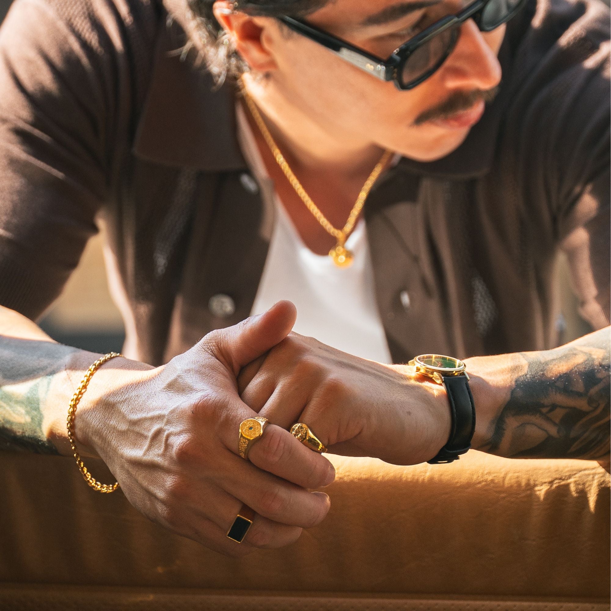 A close-up lifestyle shot of a man wearing multiple gold jewelry pieces, including a gold chain necklace, a gold bracelet, a gold watch with a black strap, and several gold rings, while wearing a dark brown shirt and sunglasses.