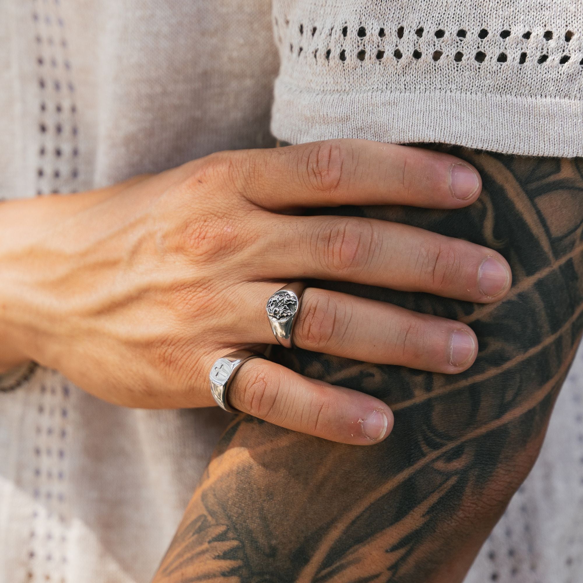 A close-up lifestyle shot of a person's hand resting on their tattooed arm, wearing two silver rings: one with a textured, hammered circular face and another with a white inlay.