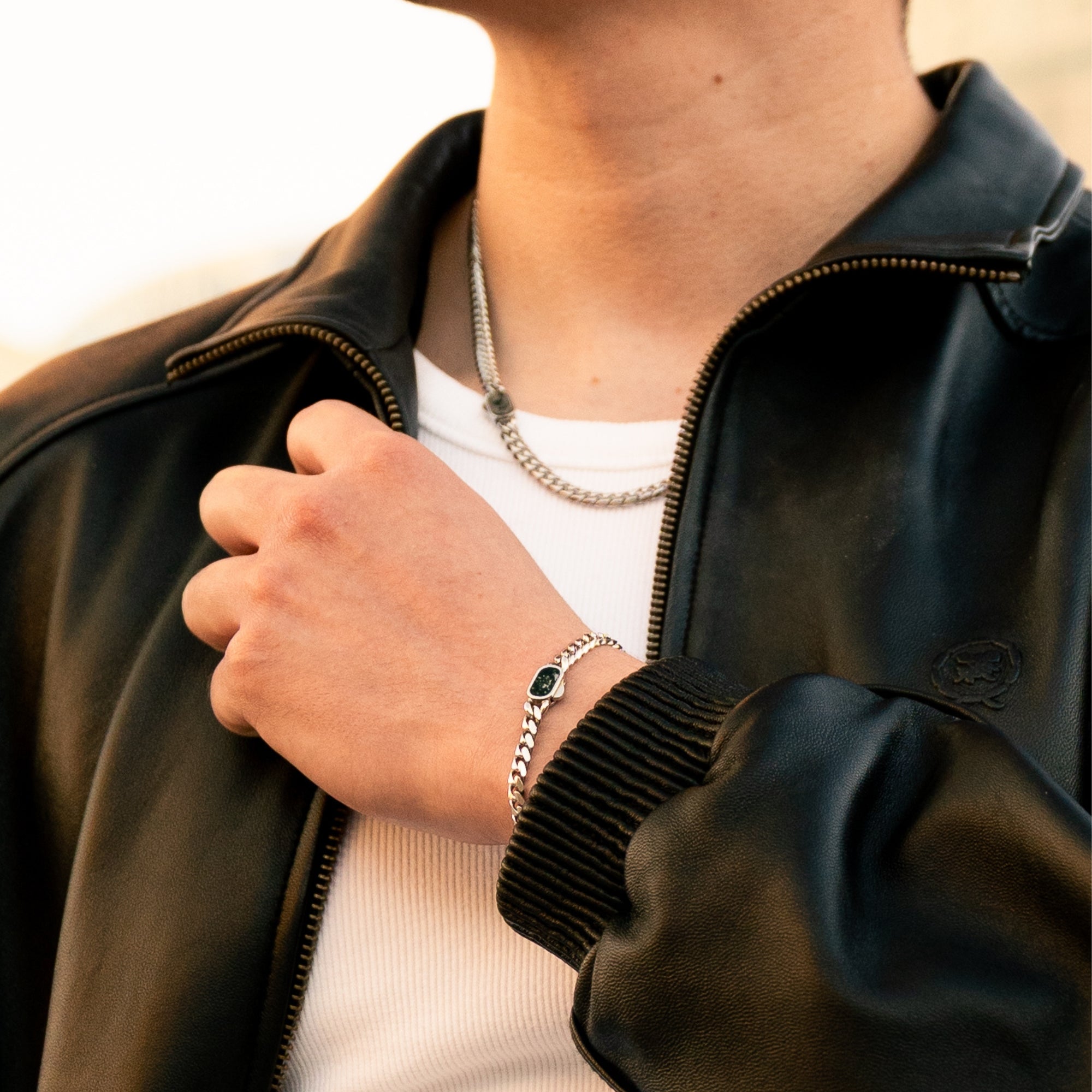 Person wearing a black leather jacket with a hand adjusting the zipper, wearing a silver chain necklace and bracelet.