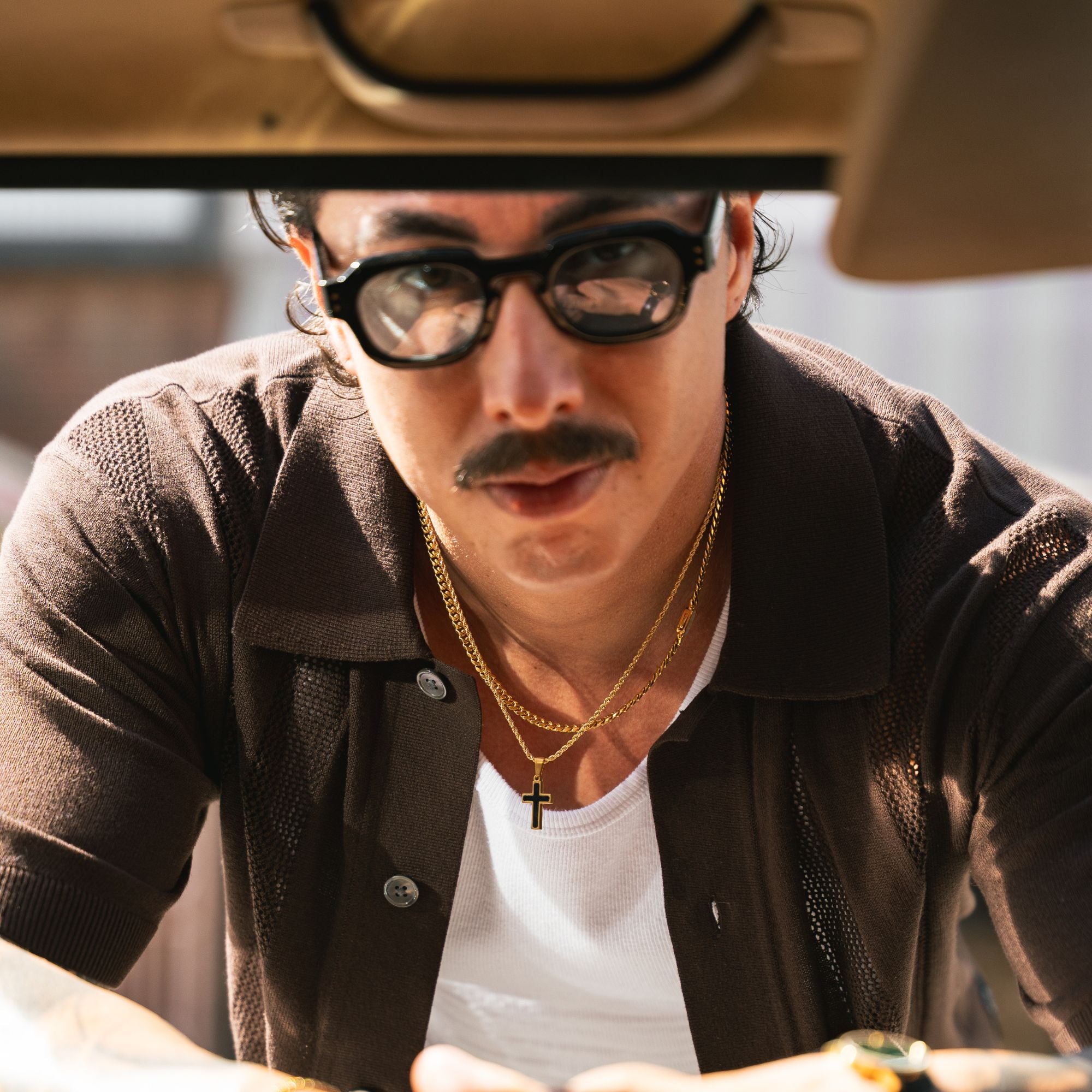 A close-up lifestyle shot of a man wearing black-rimmed glasses, a brown textured button-up shirt over a white t-shirt, and gold necklaces, looking down inside a vehicle.