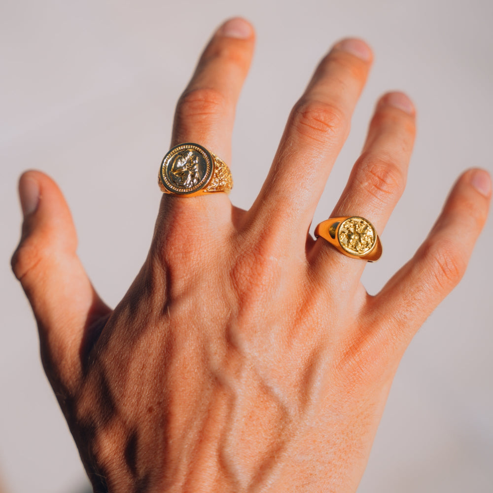 Hand wearing two gold rings with circular designs on a neutral background