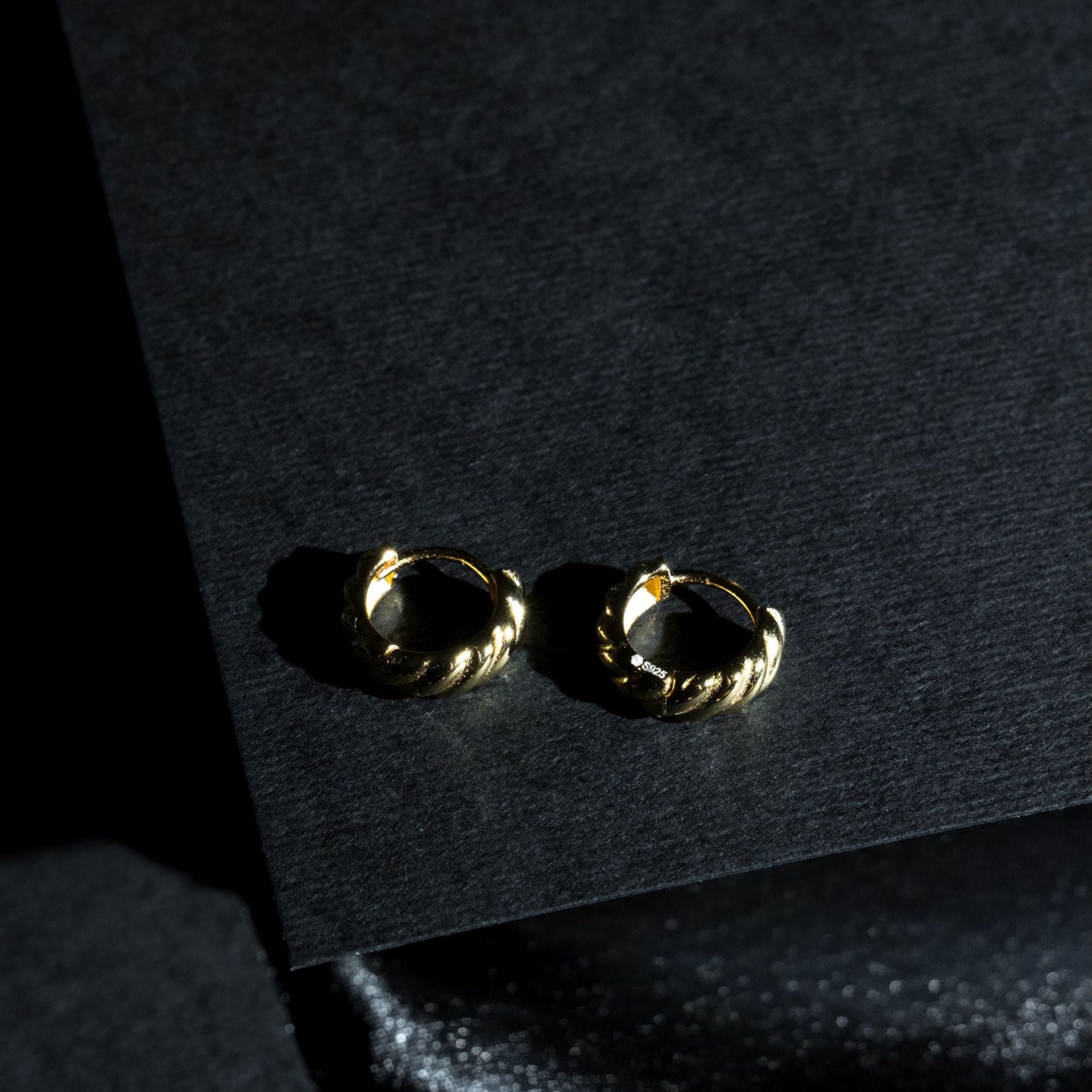 A pair of small gold huggie hoop earrings with a twisted rope texture, resting on a dark textured surface.