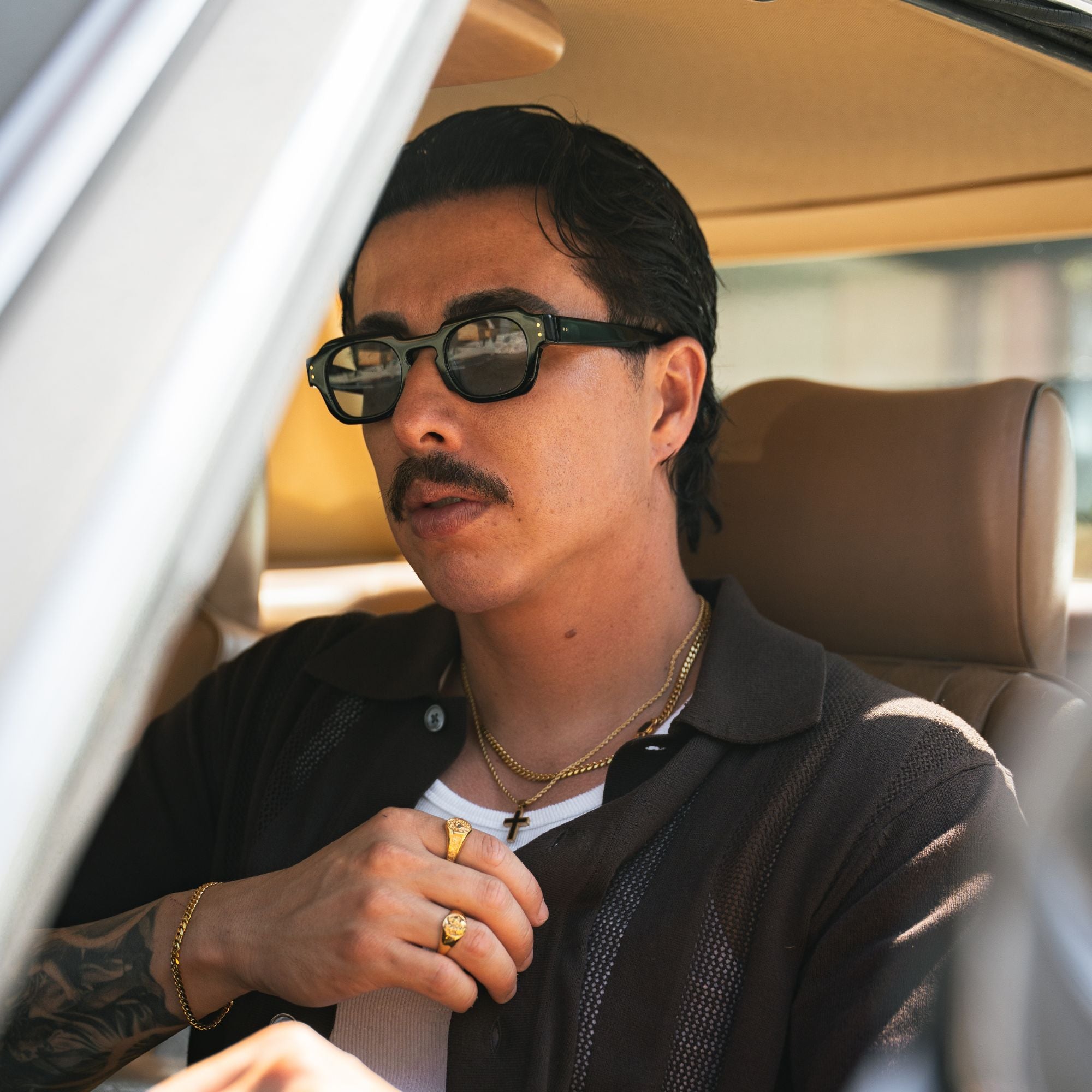 A man wearing black sunglasses with a gold cross pendant and gold rings sits inside a car with tan leather seats.