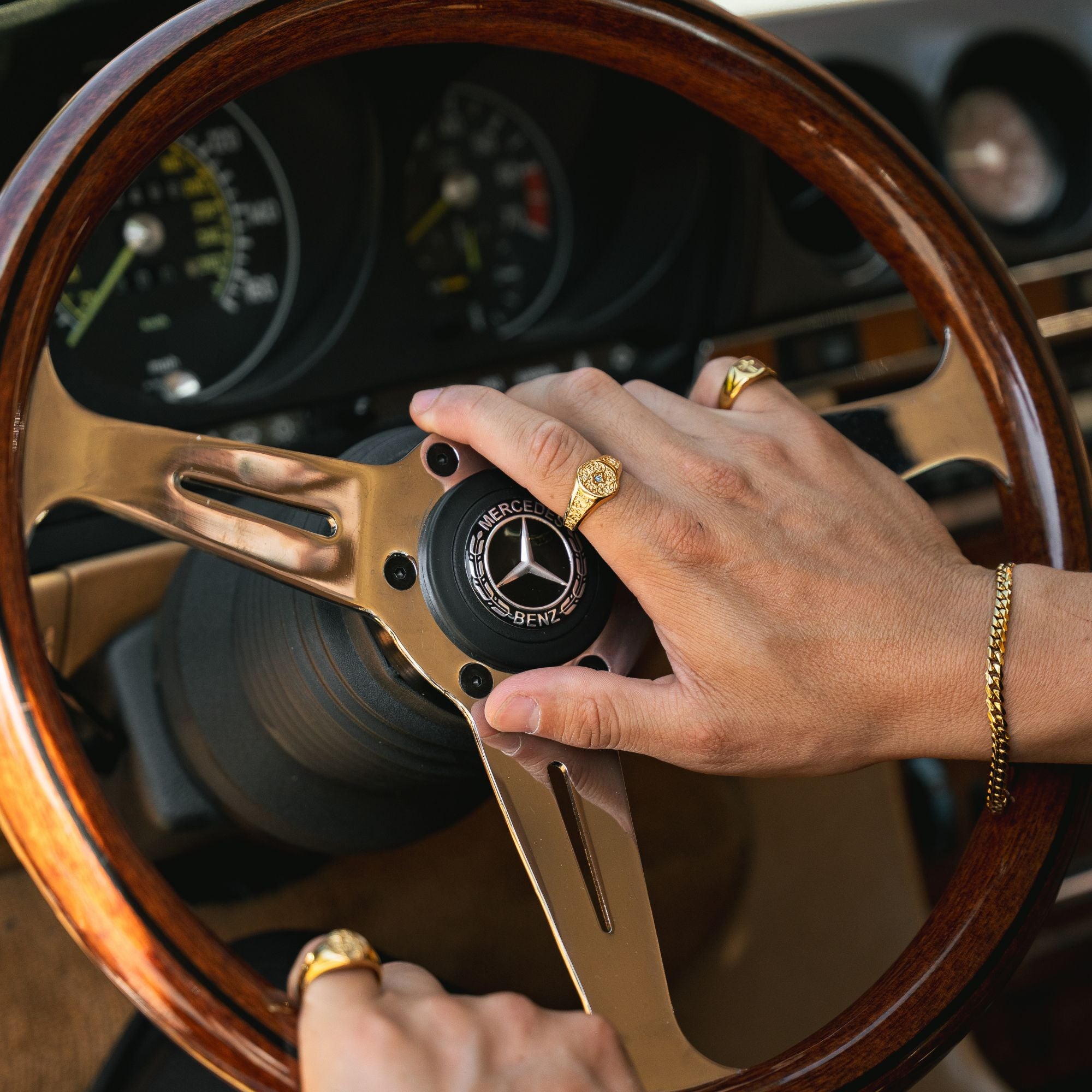 A close-up of a person's hands on a vintage-style wooden steering wheel with a polished gold center cap featuring the Mercedes-Benz logo. The person is wearing gold rings and a gold chain bracelet inside a car cabin with dashboard gauges visible in the background.