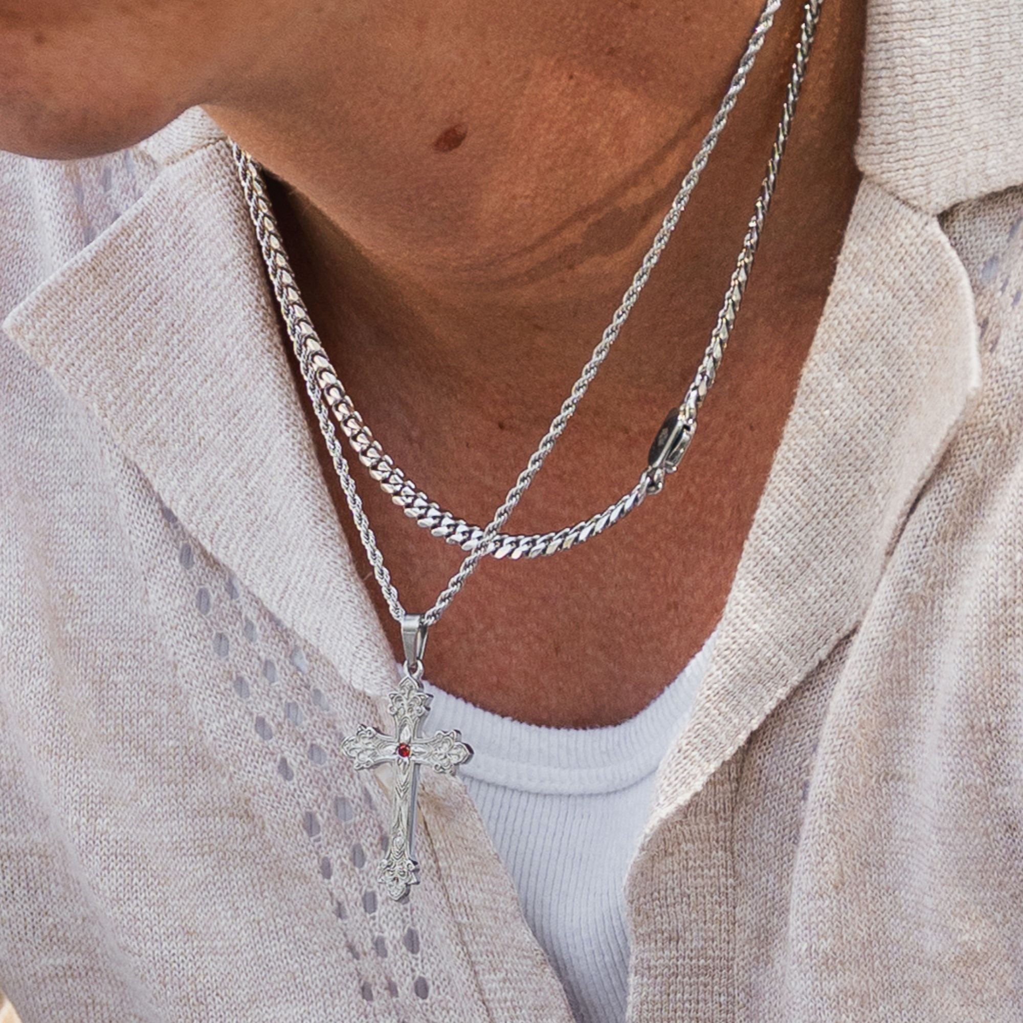 A close-up of a person wearing two layered silver necklaces, one curb chain and one twisted rope chain, both ending in a cross pendant. The cross pendant features a small red gemstone at its center.