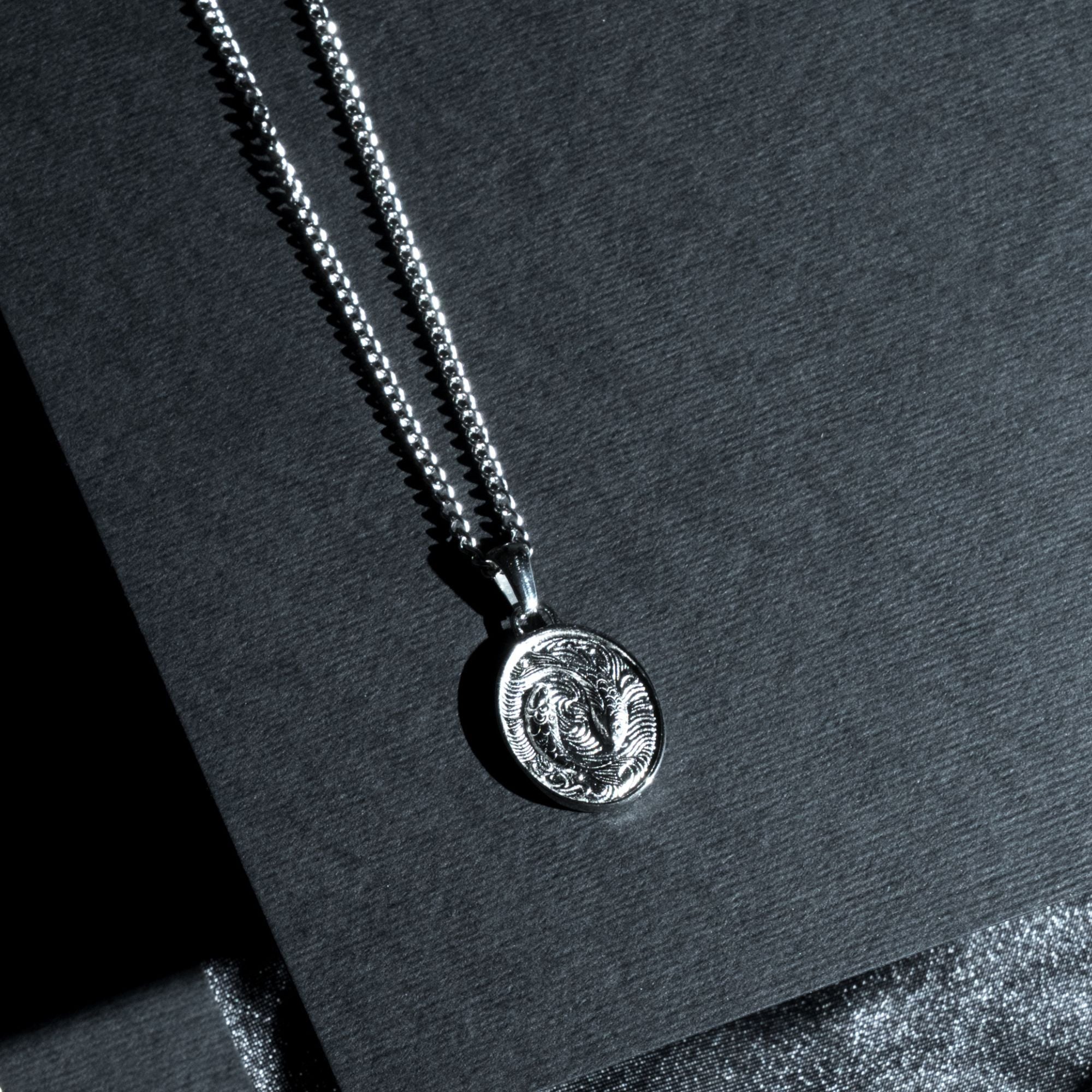 A silver chain necklace with a circular coin-style pendant featuring an engraved ram design, displayed on a dark textured surface.