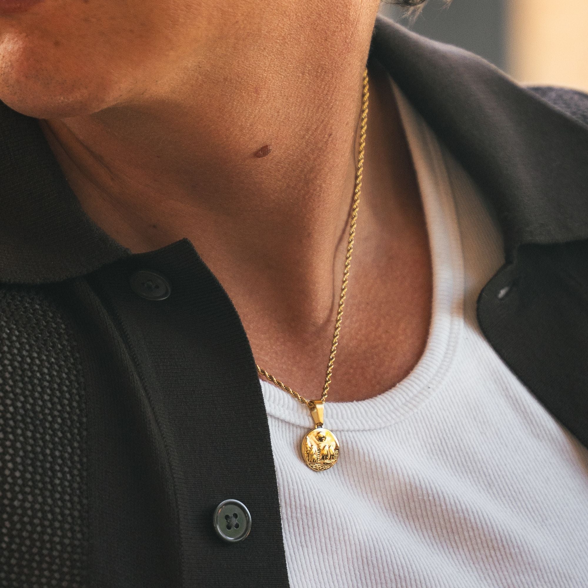 A close-up of a person wearing a gold pendant necklace with a textured circular medallion, styled over a white ribbed t-shirt and a black button-up shirt.