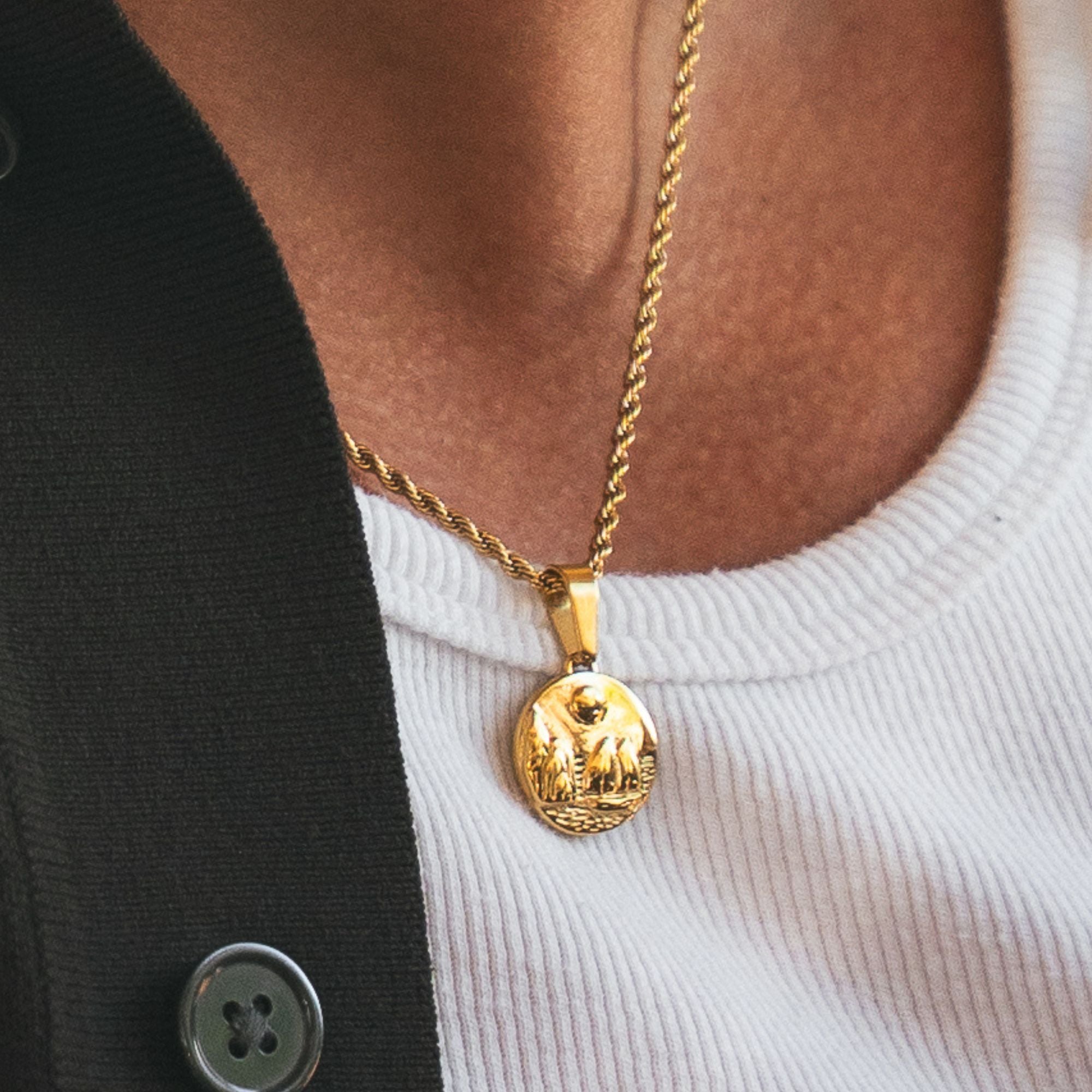 A close-up of a person wearing a gold pendant necklace with a circular engraved medallion on a twisted chain, styled over a white ribbed top and a black cardigan.