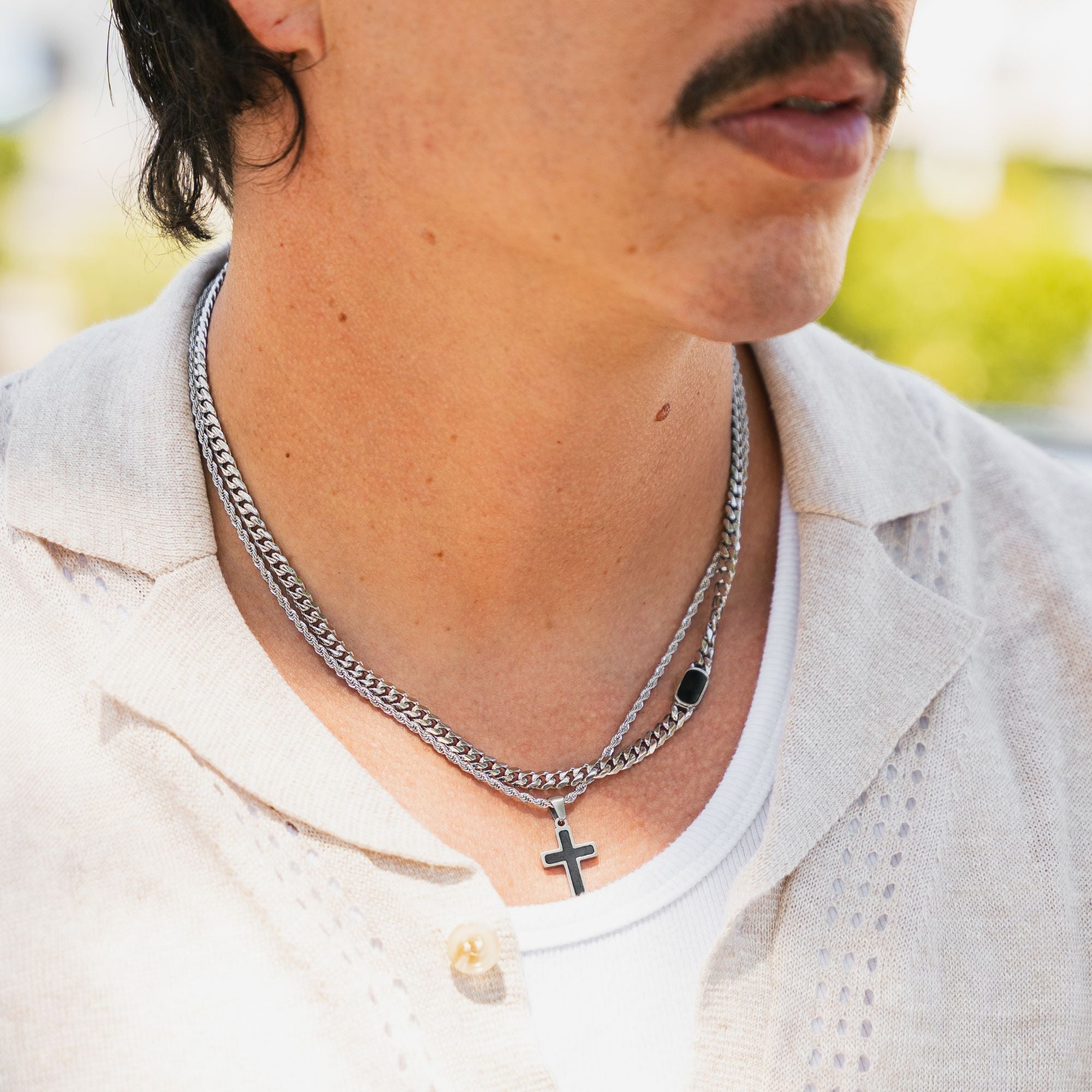 A close-up lifestyle shot of a man wearing a double-layered silver chain necklace. The top layer features a classic curb chain, while the bottom layer is a smoother cable chain with a small black cross pendant and a black rectangular charm.